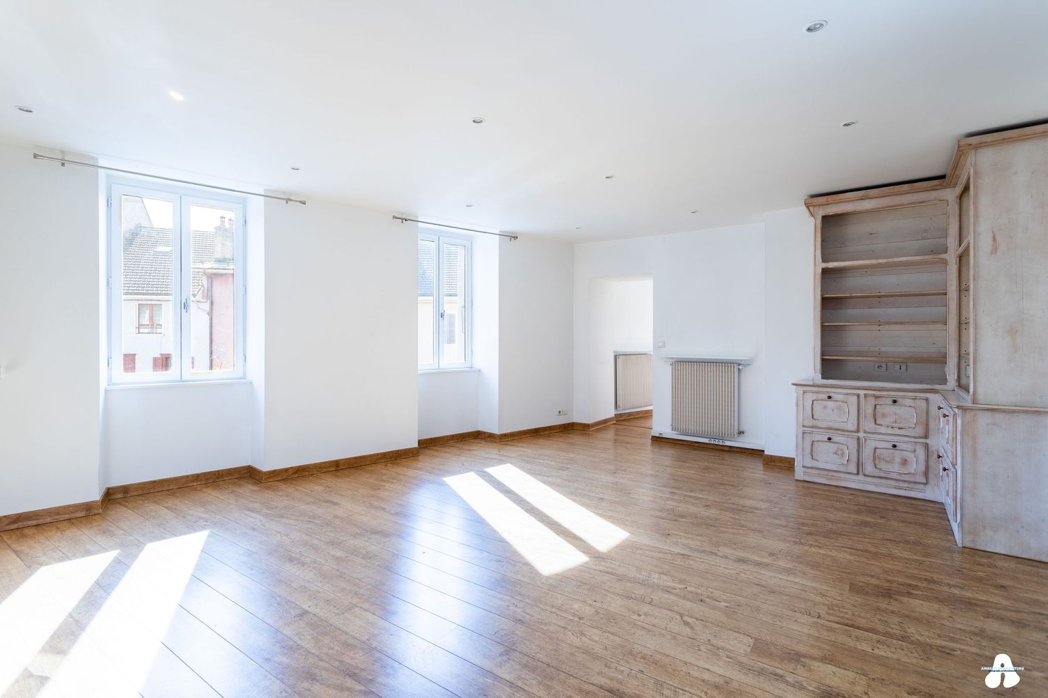 Bright empty living room with hardwood floors, white walls, built-in bookshelf and large windows letting in natural light.