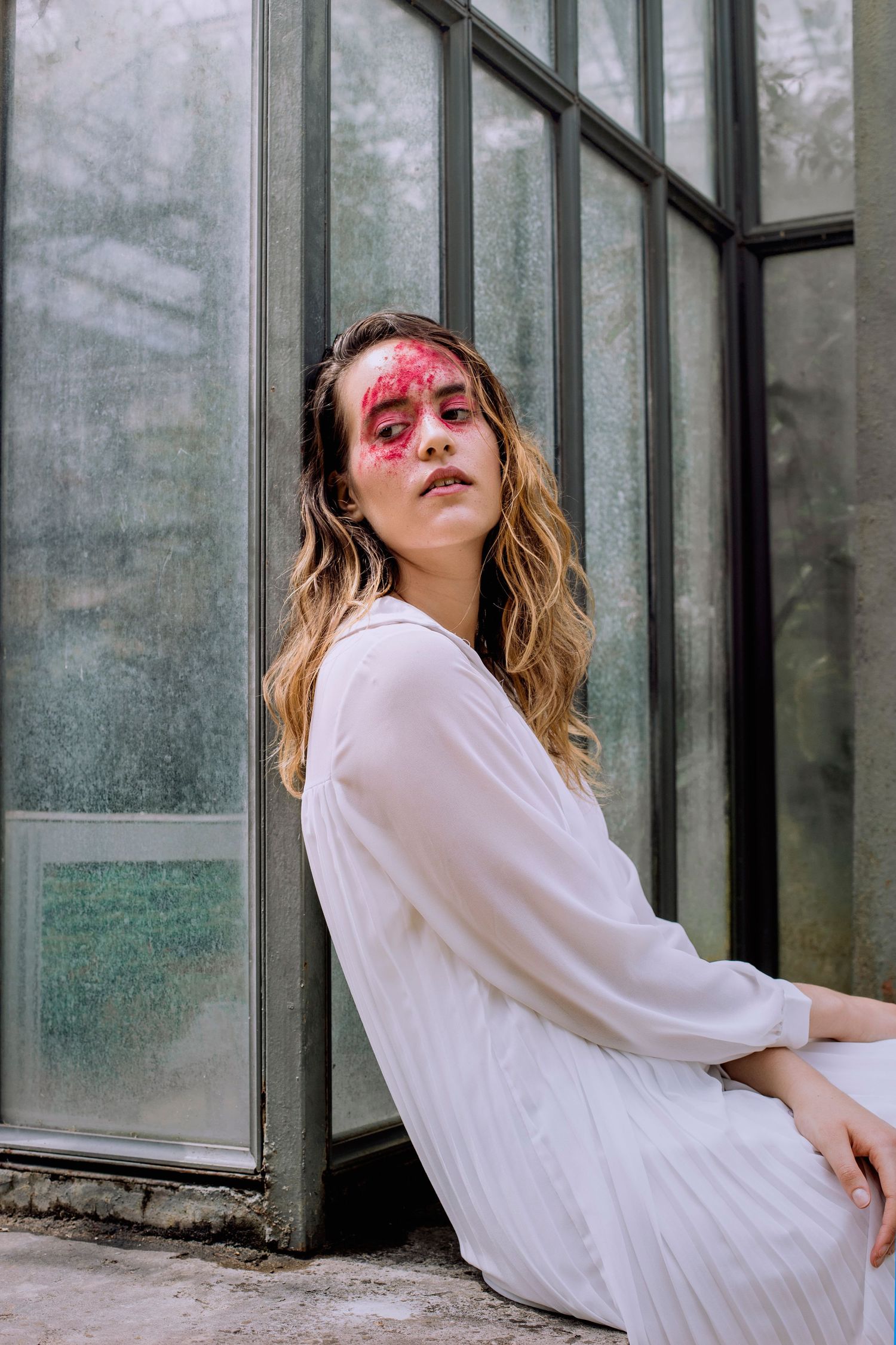 Woman in white dress with red face art leaning against glass wall.