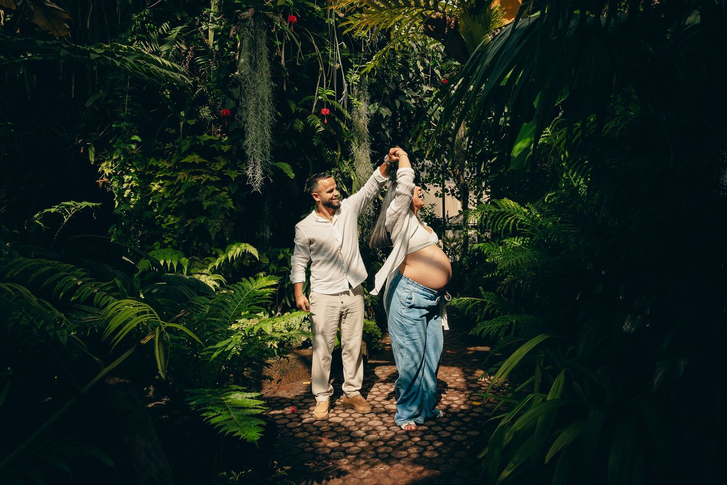 Couple dancing joyfully in a lush tropical garden, woman showing pregnant belly.
