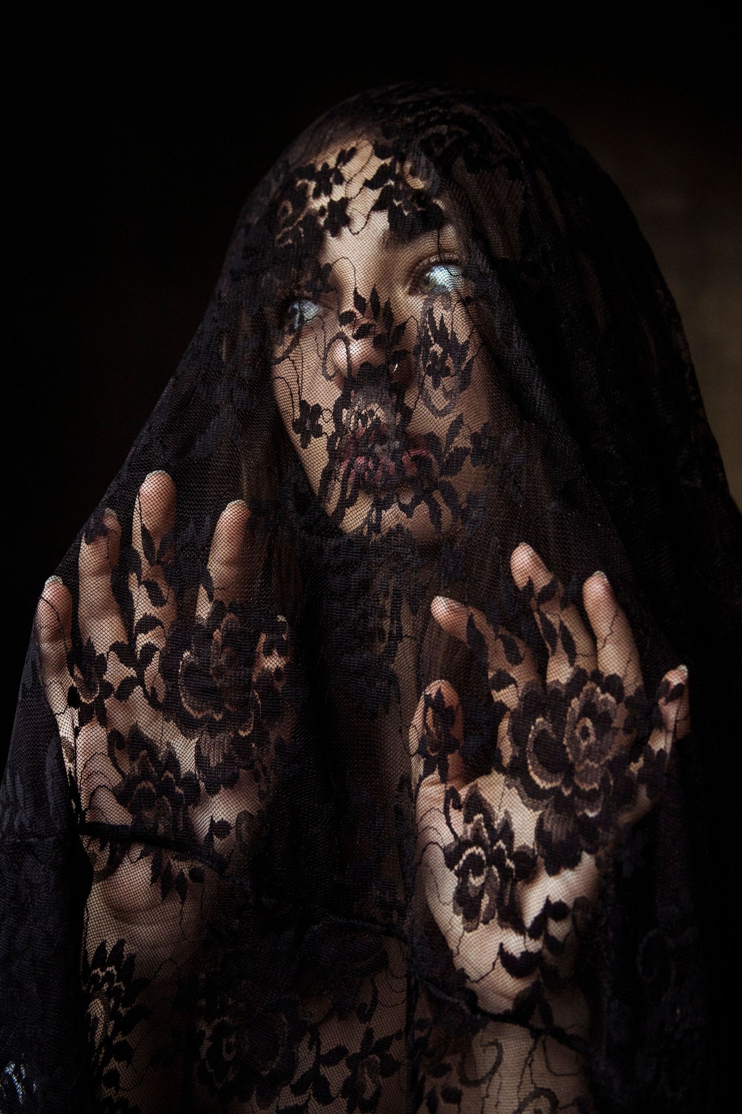 Woman with striking blue eyes covered by black floral lace veil with hands raised in dramatic portrait.