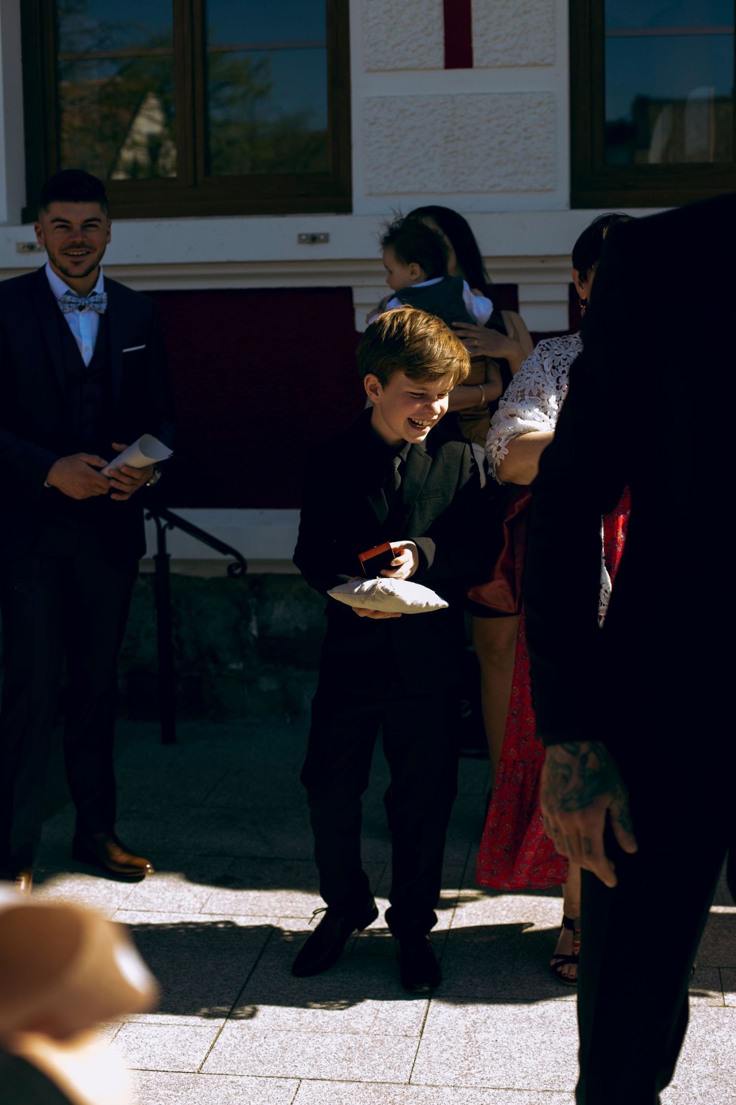 Young boy in black suit smiles while holding ring pillow outside at sunny wedding celebration.