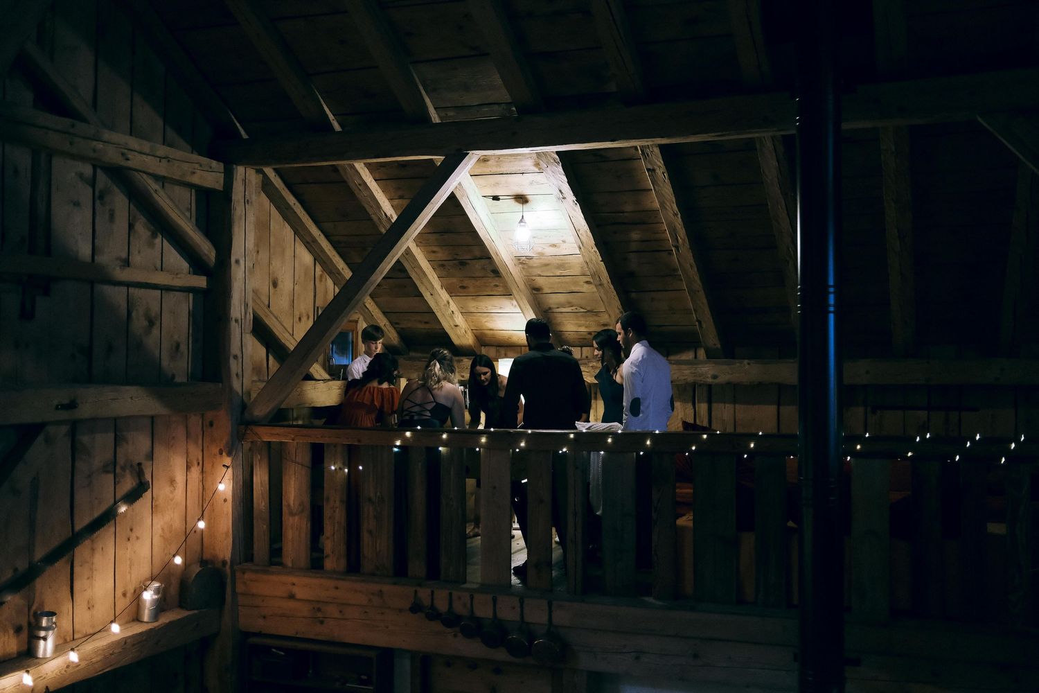 Guests gather on dimly lit wooden barn loft balcony decorated with string lights.