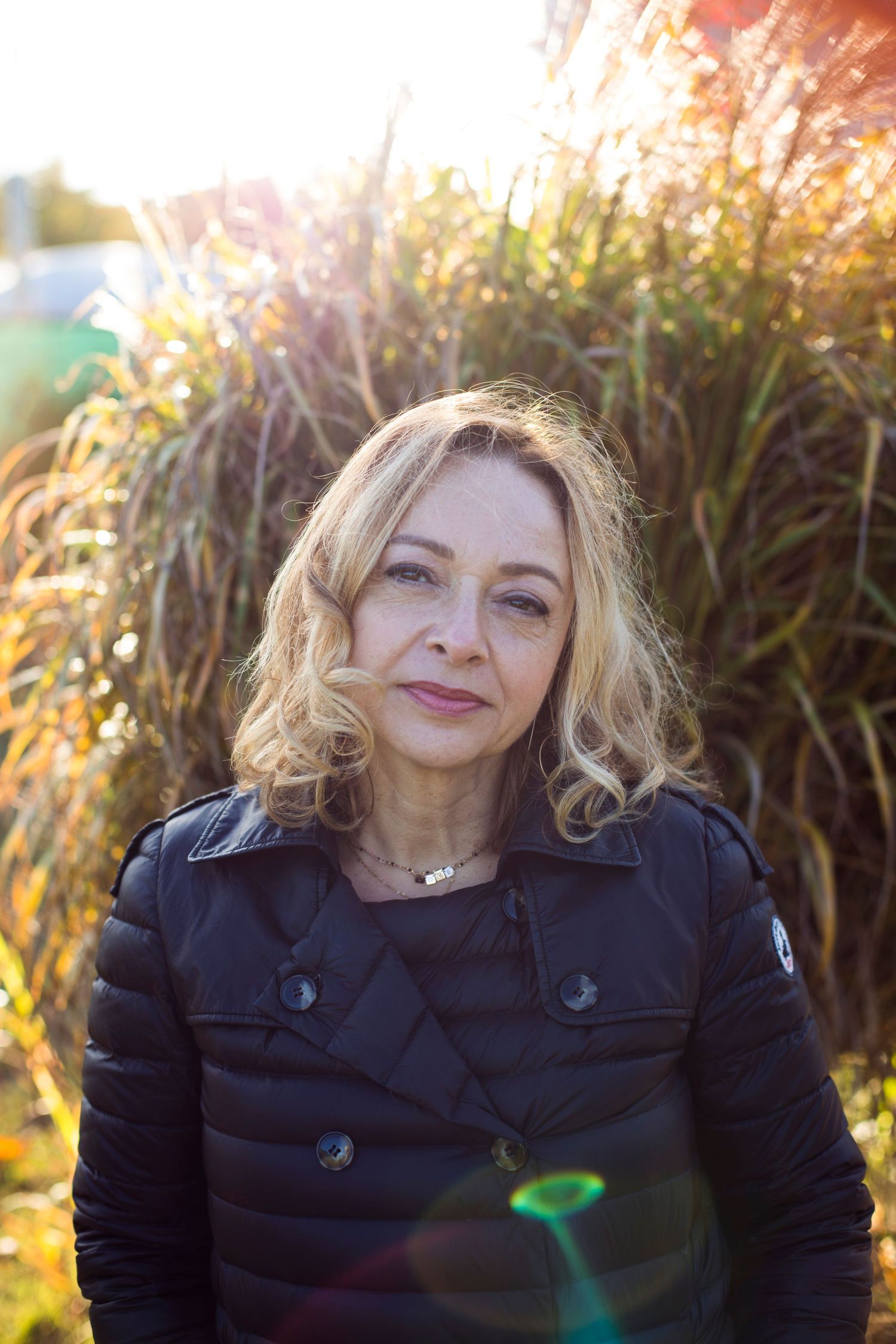 Portrait of a middle-aged blonde woman in a black jacket standing in sunlit autumn grass outdoors.