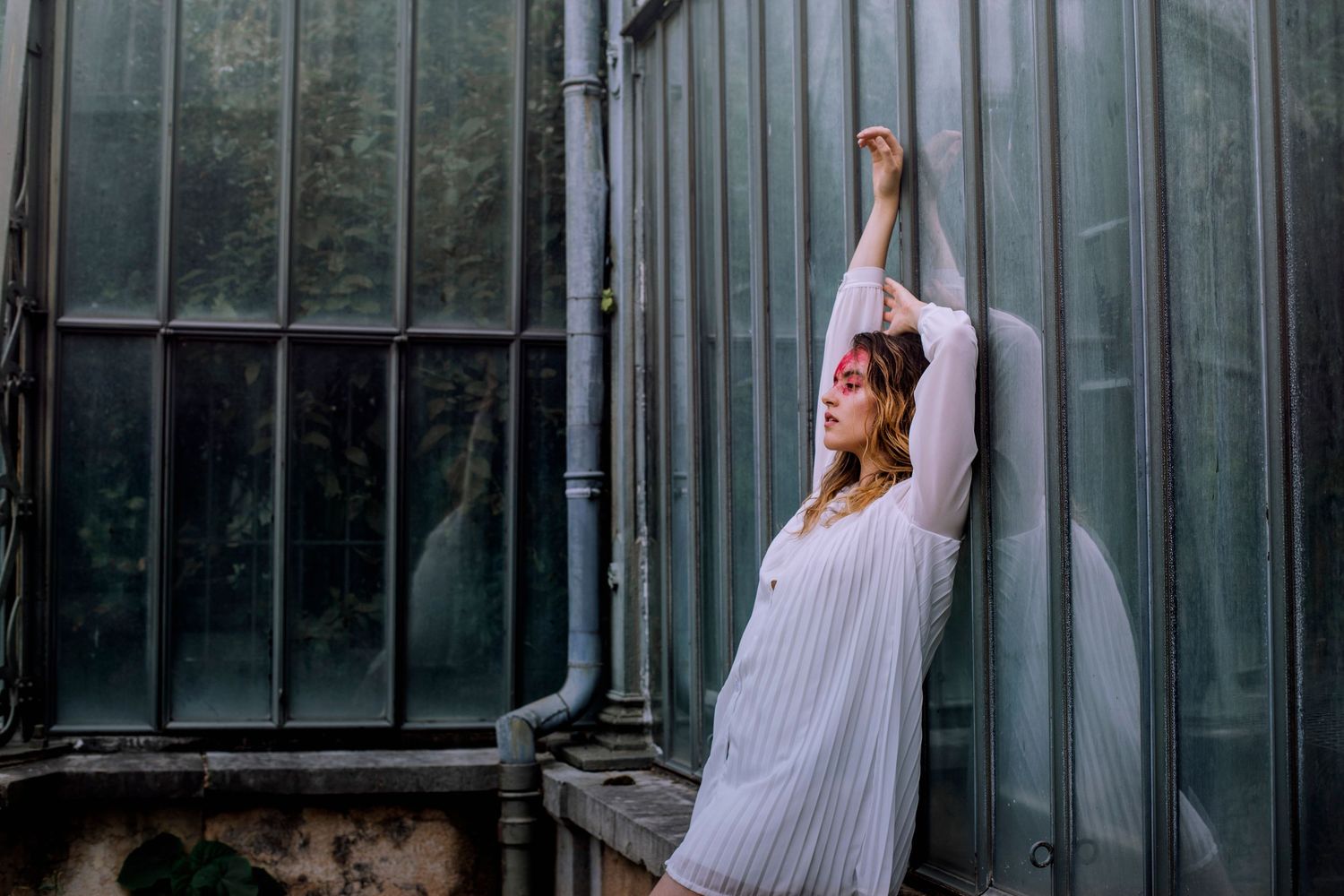Woman in white dress stretching arms beside a vintage greenhouse wall.