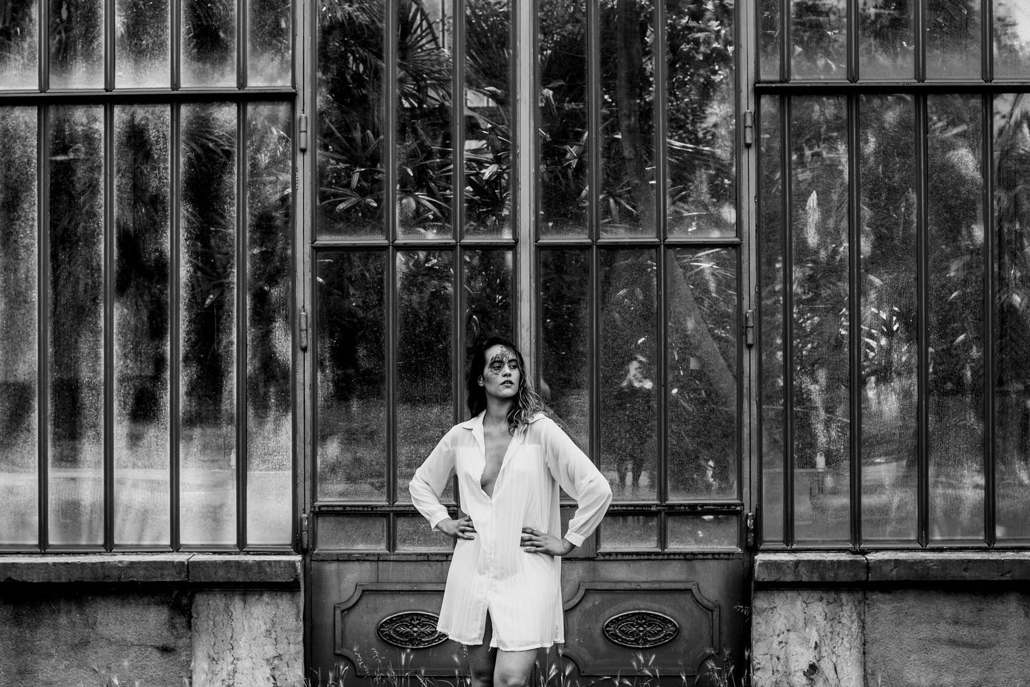 Black and white full-length photo of a woman in a white outfit posing before large ornate greenhouse windows.