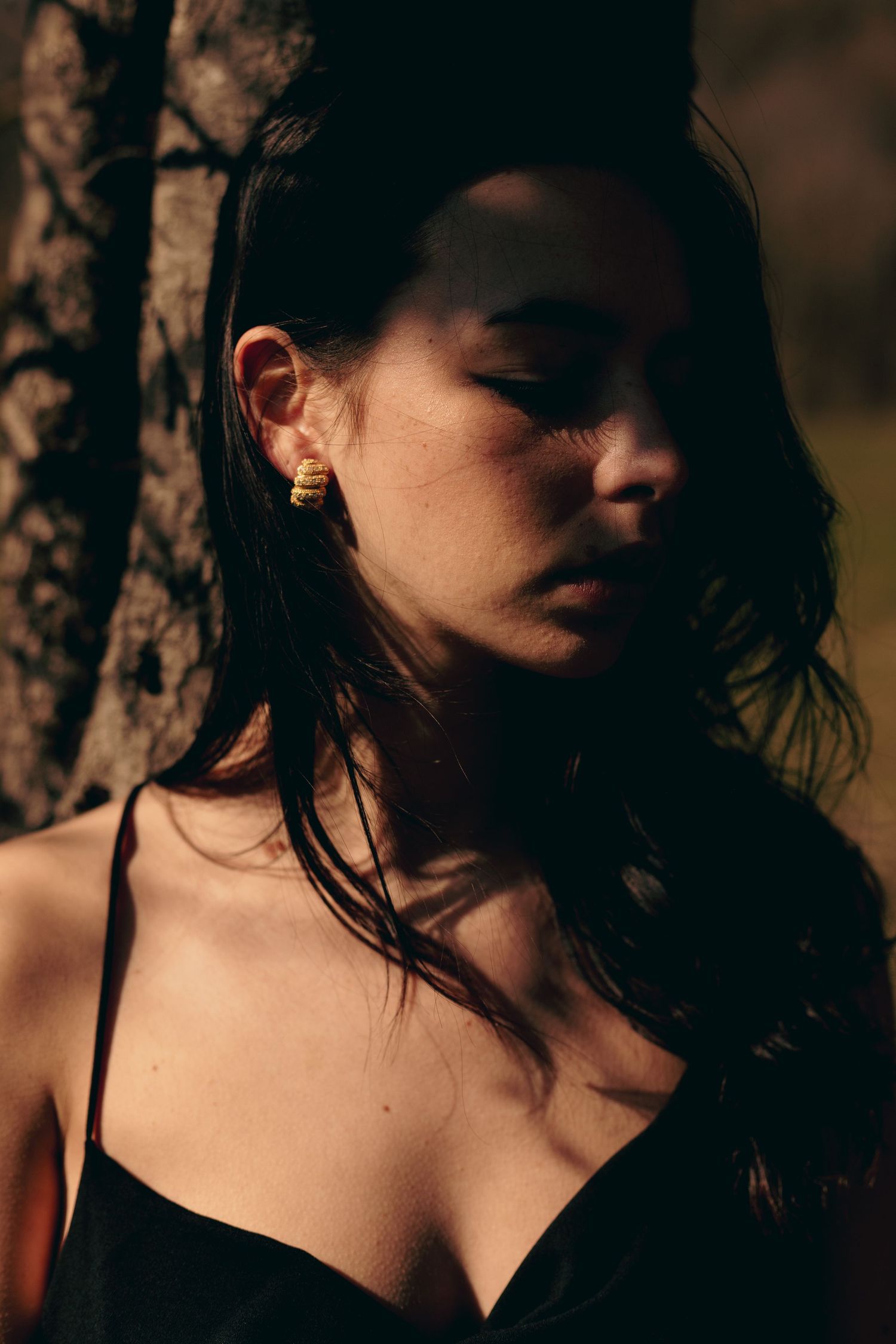 Young woman with dark hair and gold earring poses eyes closed near tree in warm sunlight.