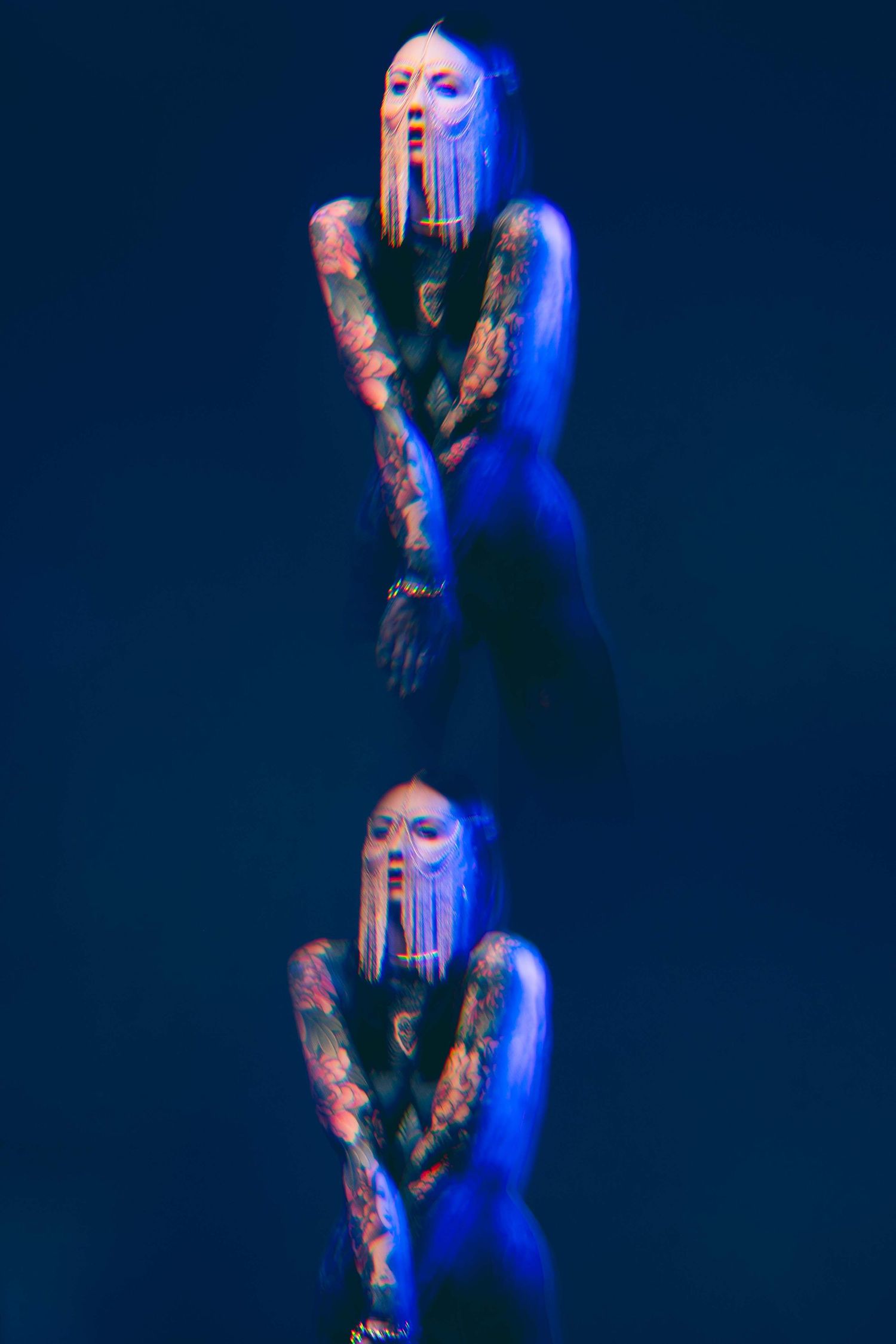 Double exposure portrait of tattooed woman with chain face jewelry under deep blue studio lighting.