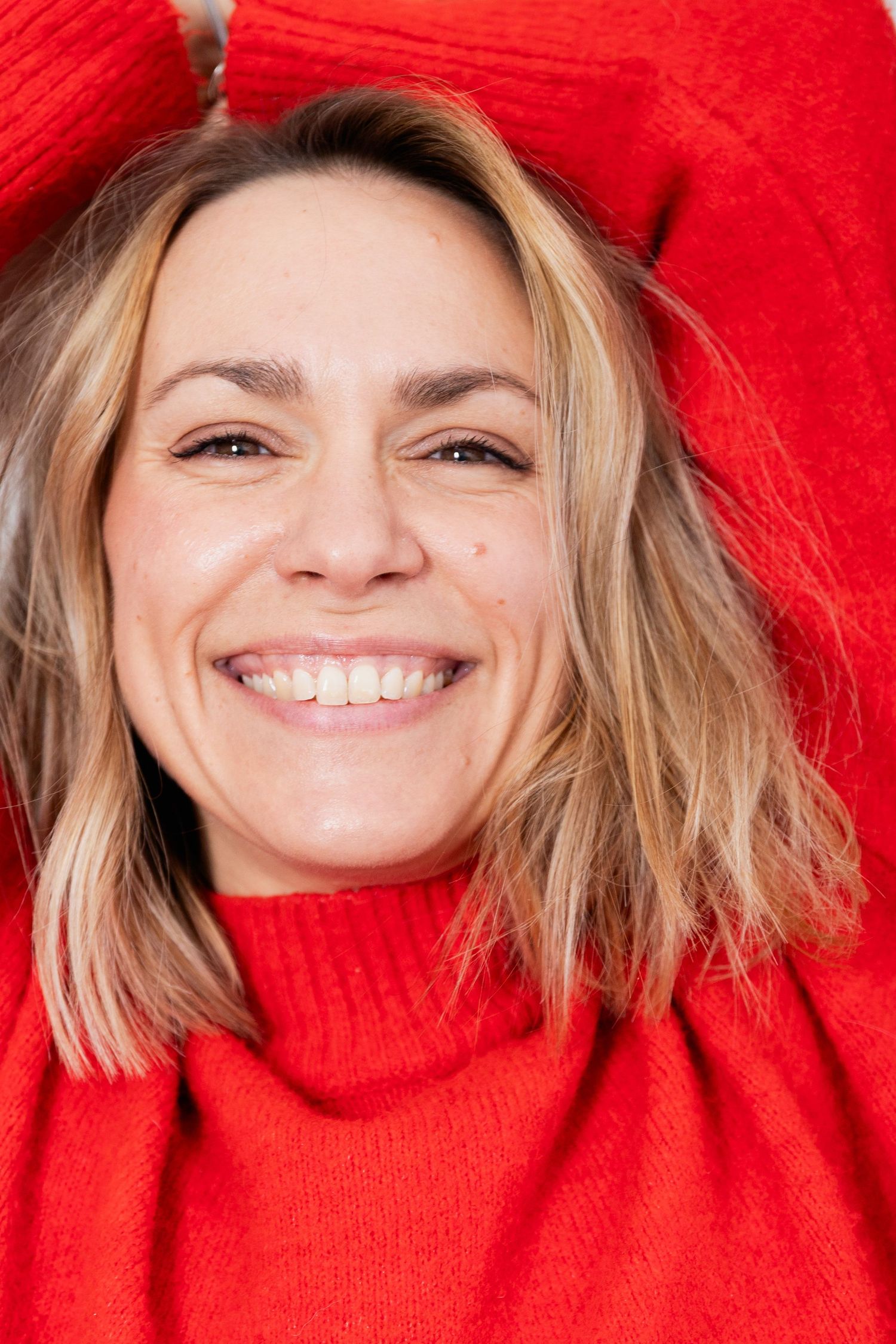Smiling blonde woman in red turtleneck sweater, close-up portrait with hands behind head.