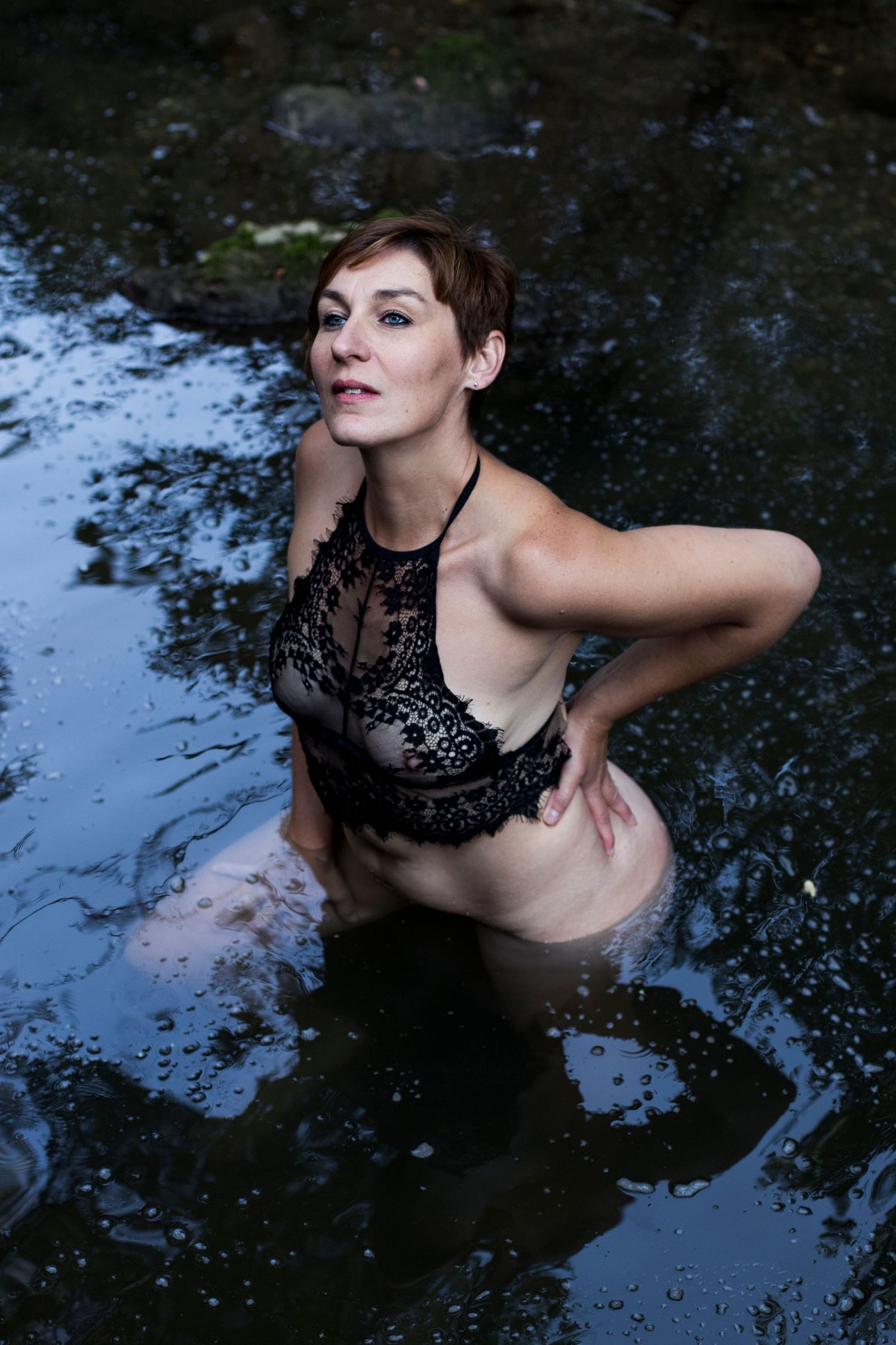 Woman in black lace bralette posing in a dark rocky stream.