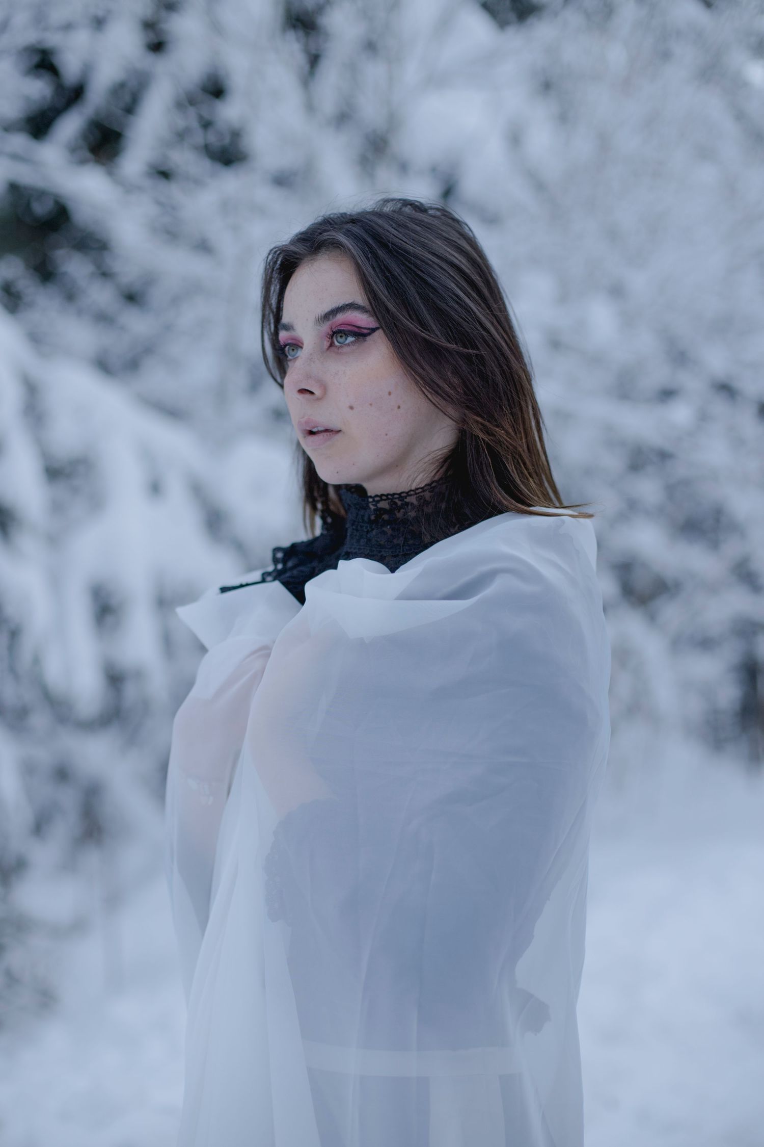 Young woman with pink eye makeup wrapped in white sheer fabric in snowy winter forest.