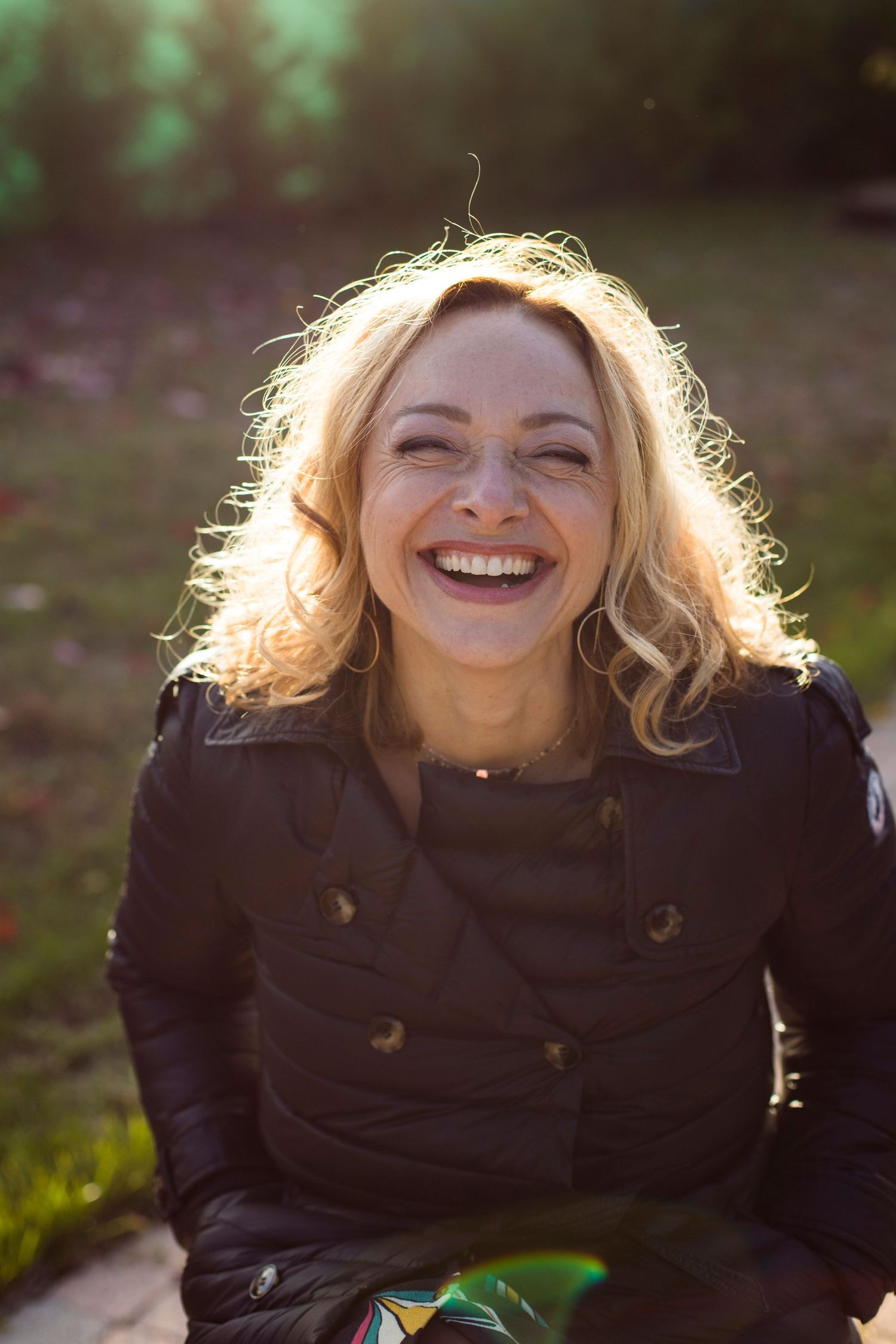Joyful blonde woman in a black jacket laughing in a sunlit outdoor park setting.