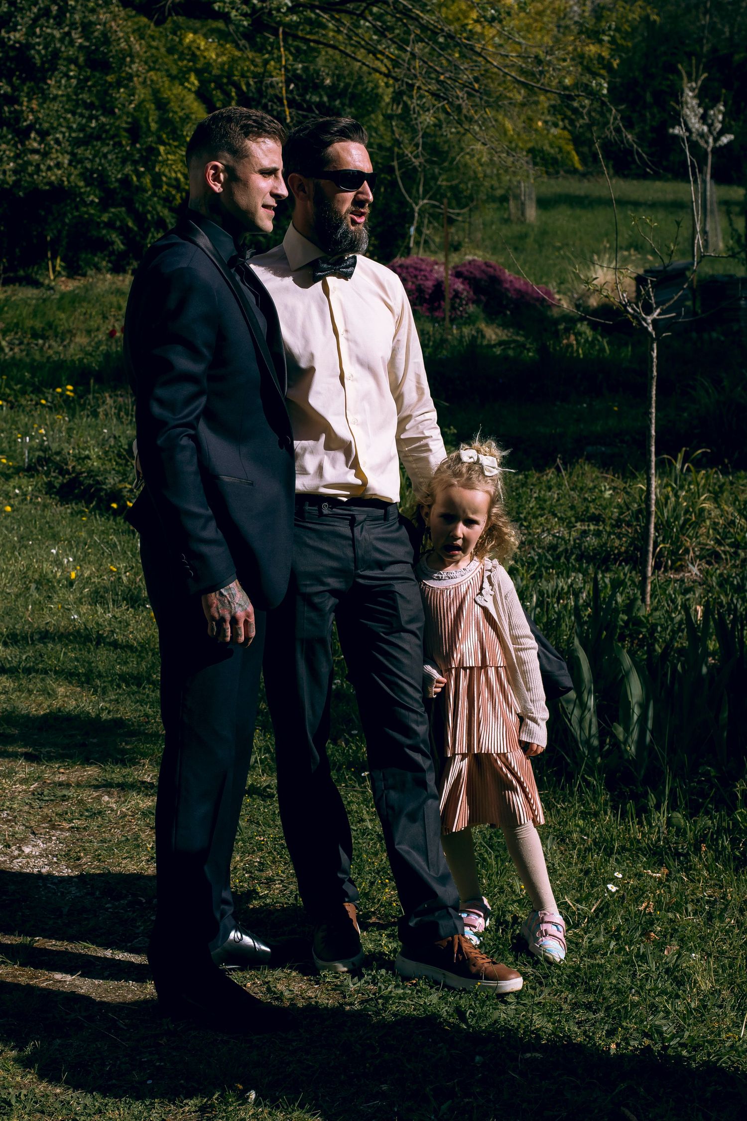 Two men in formal attire and a young girl in pink dress posing together outdoors in a sunny garden.