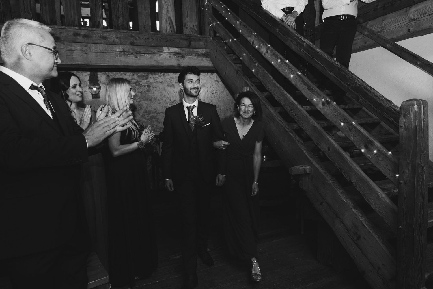 Groom descends rustic barn staircase as wedding guests applaud in black and white photo.