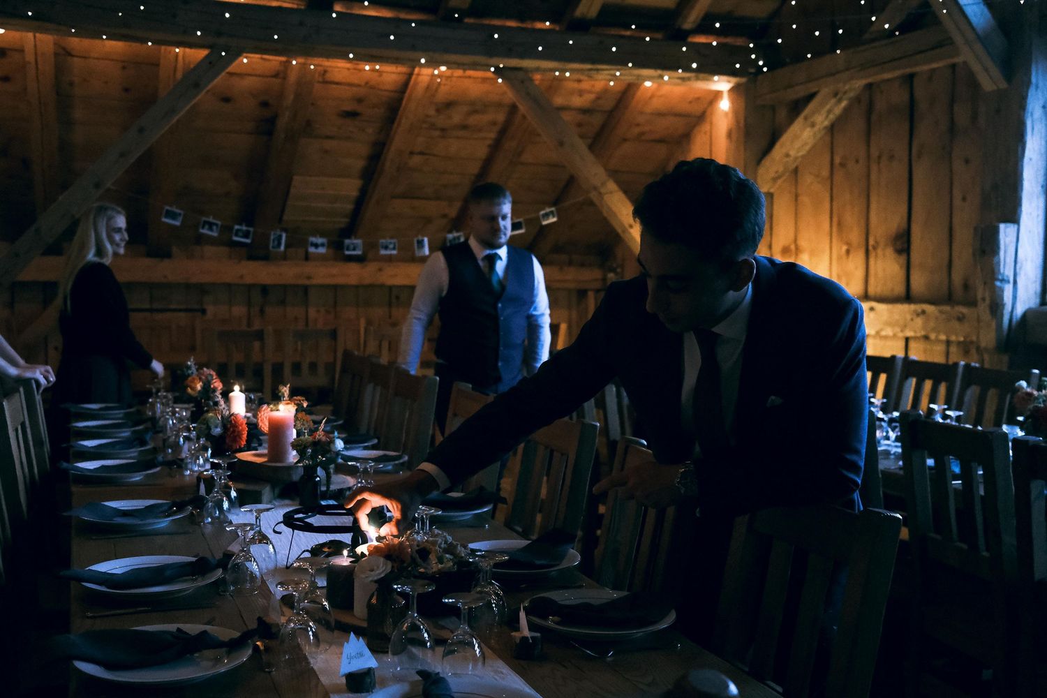 Wedding guests gather around candlelit dinner table in warmly lit rustic barn venue.