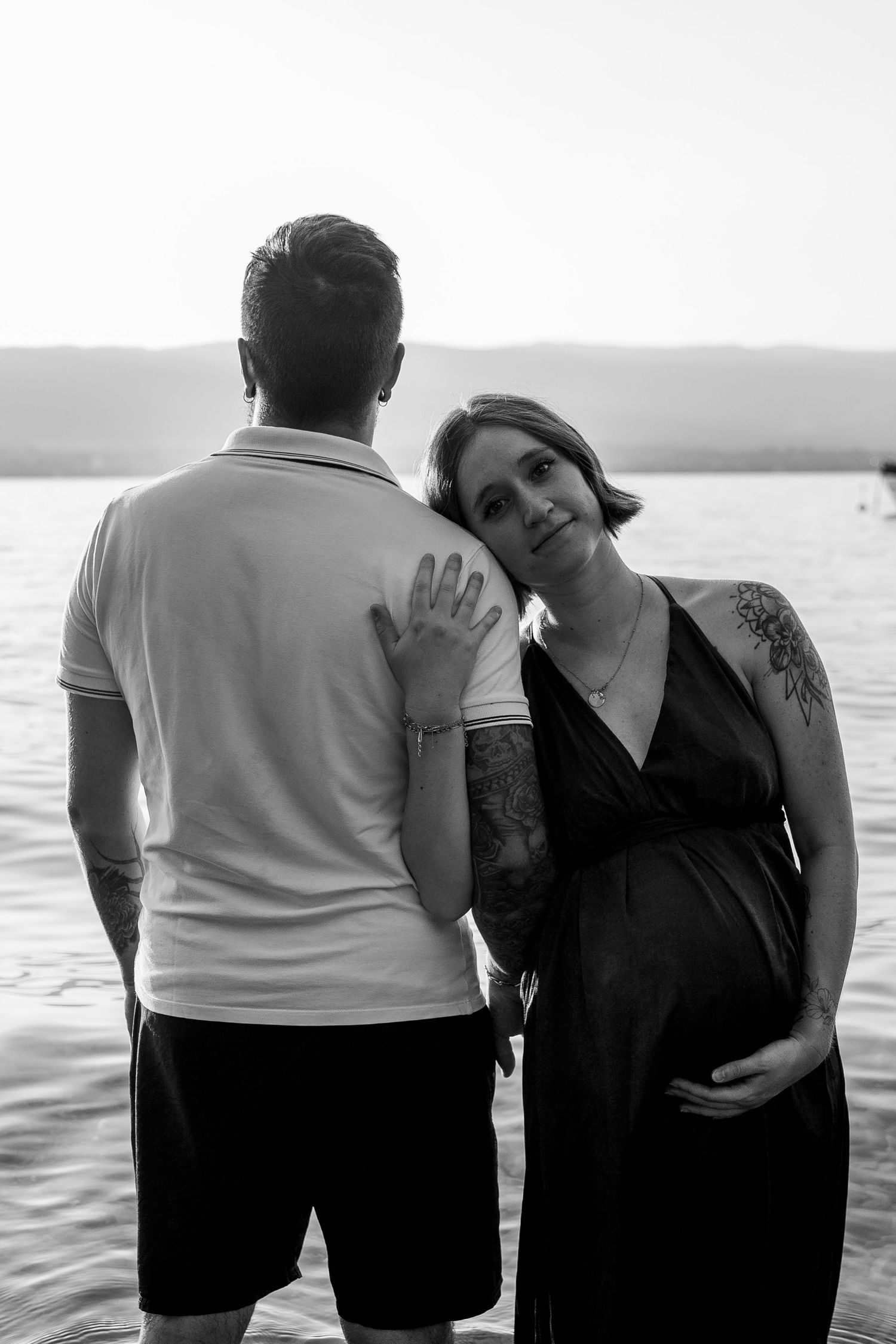 Pregnant woman in black dress leans on tattooed man by lake at sunset in black and white.