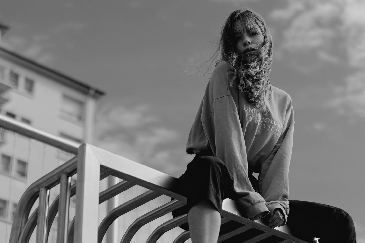 Black and white photo of young woman in oversized sweater perched on metal railings outdoors.