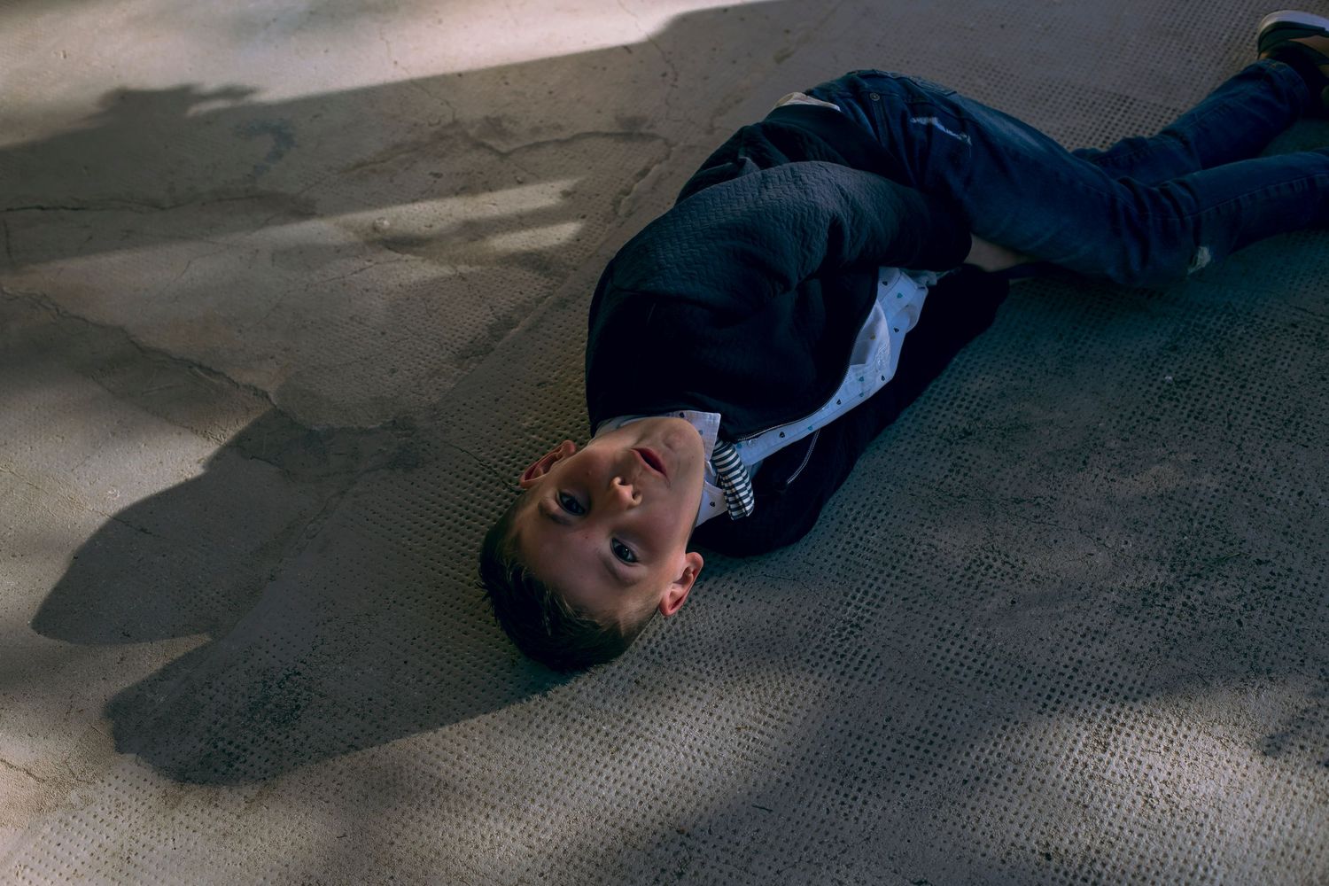 Young boy in navy suit lying on his back on pavement looking upward in natural light.