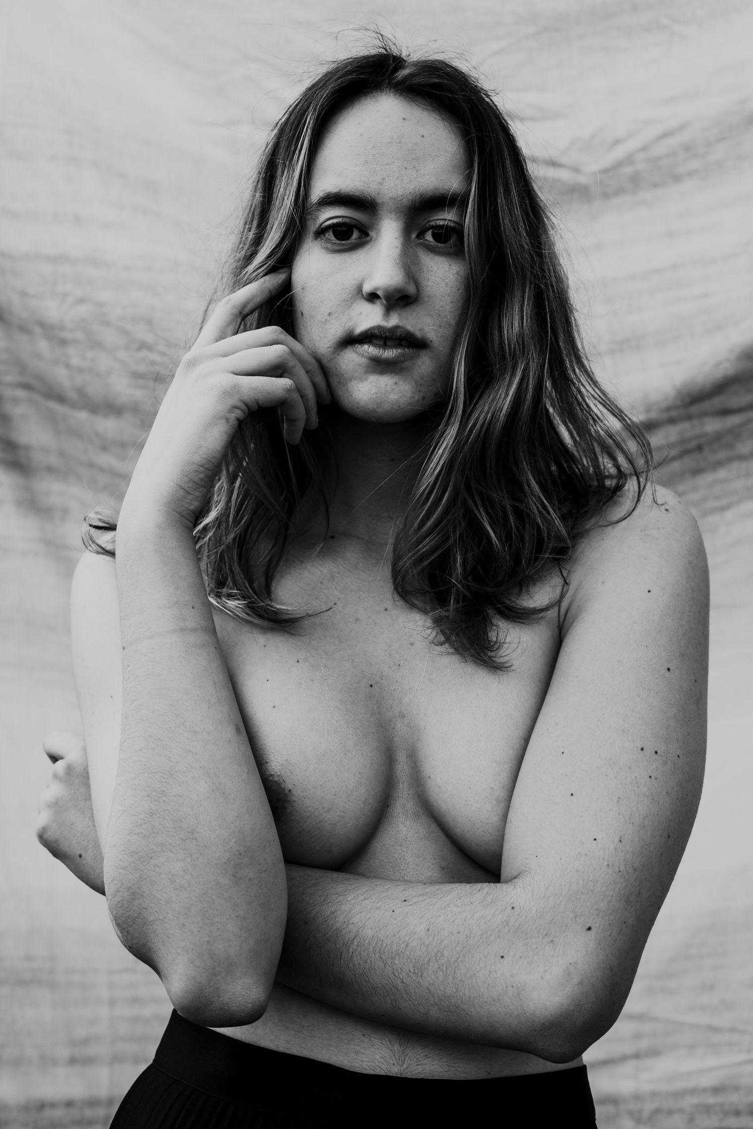 Black and white portrait of a topless woman with hand near face, arms crossed, against a fabric backdrop.