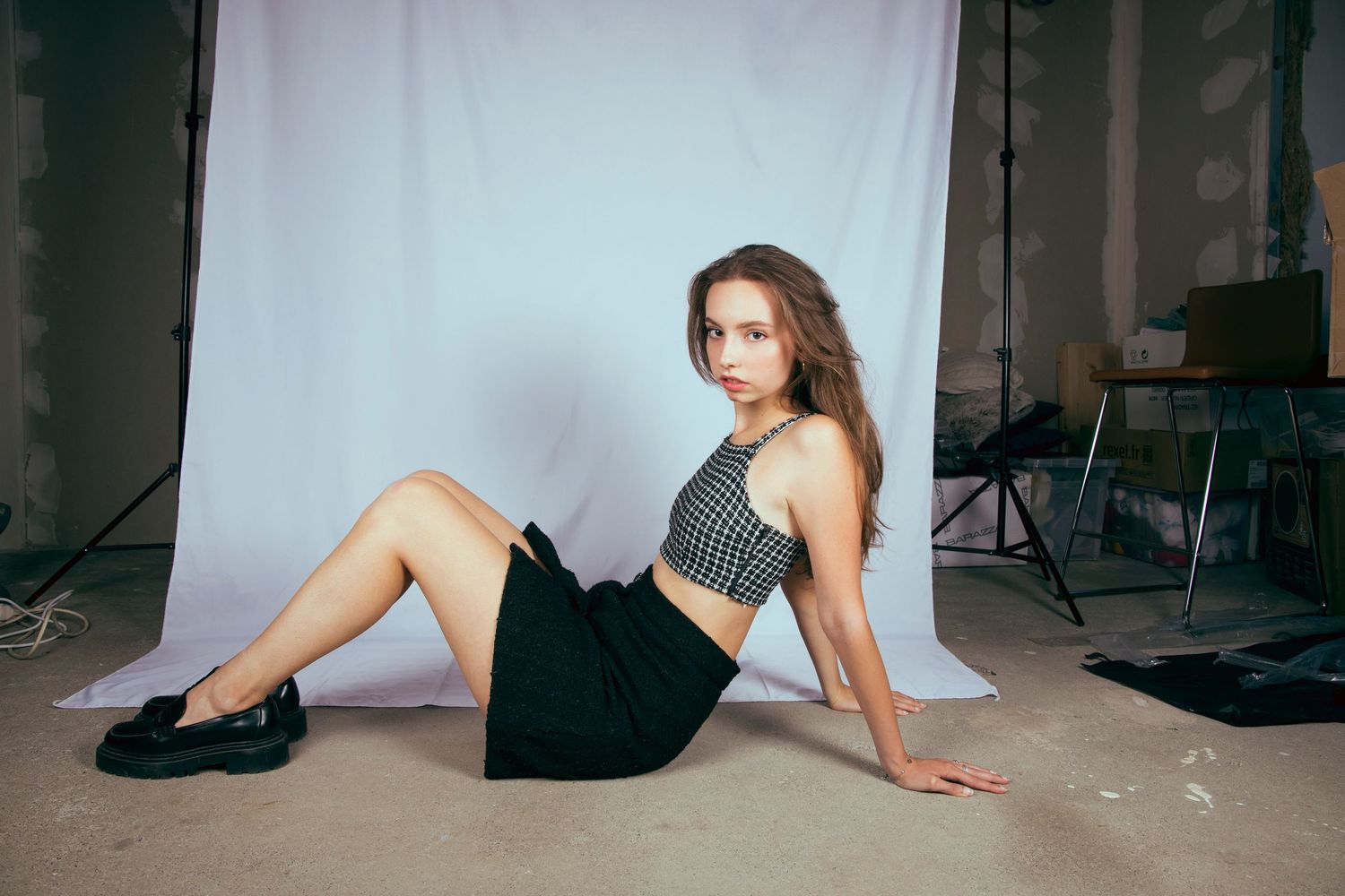 Young woman in checkered crop top and black skirt sitting on floor against white backdrop in photography studio.