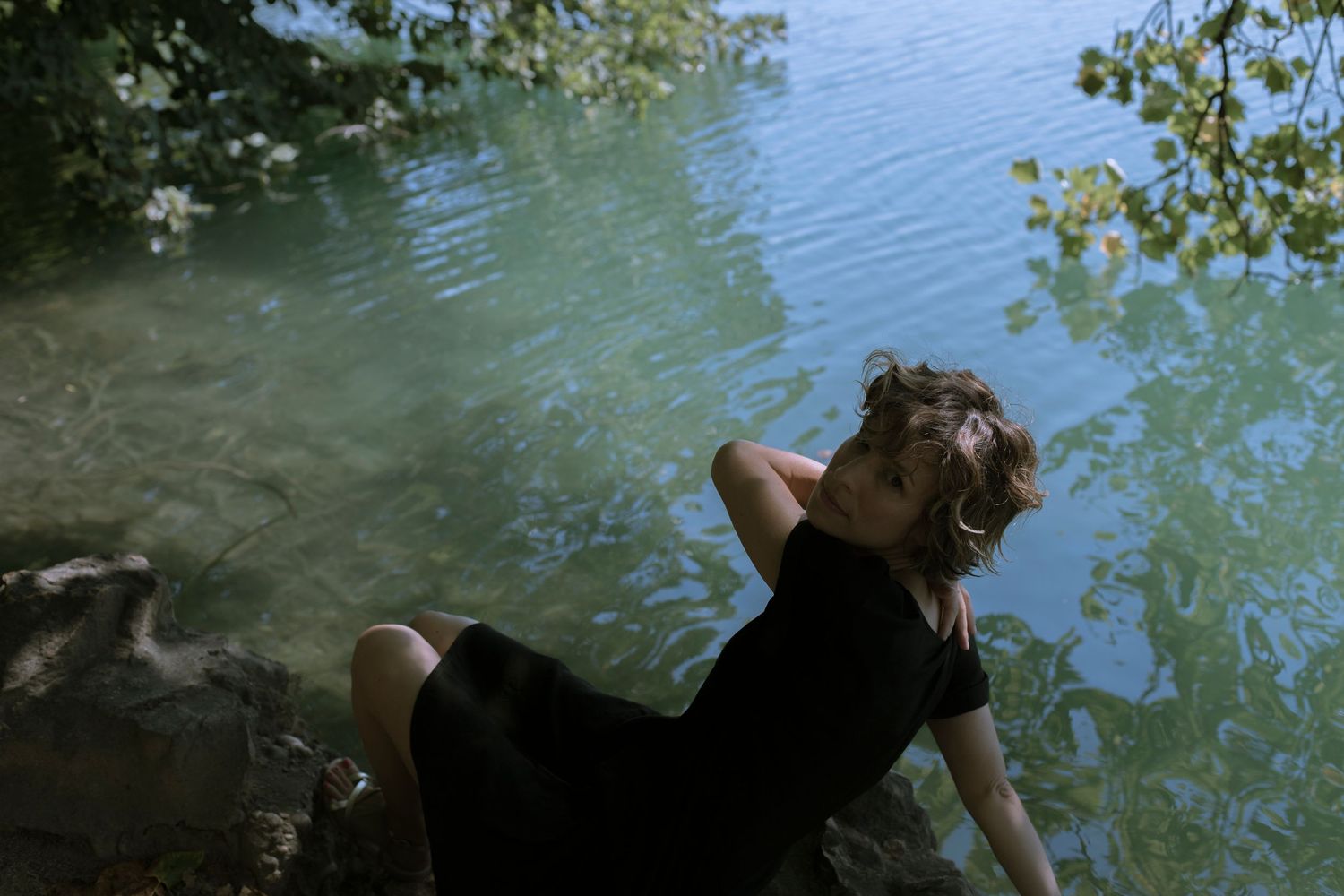 Woman in a black dress reclining on rocks by a serene green lake surrounded by lush trees.