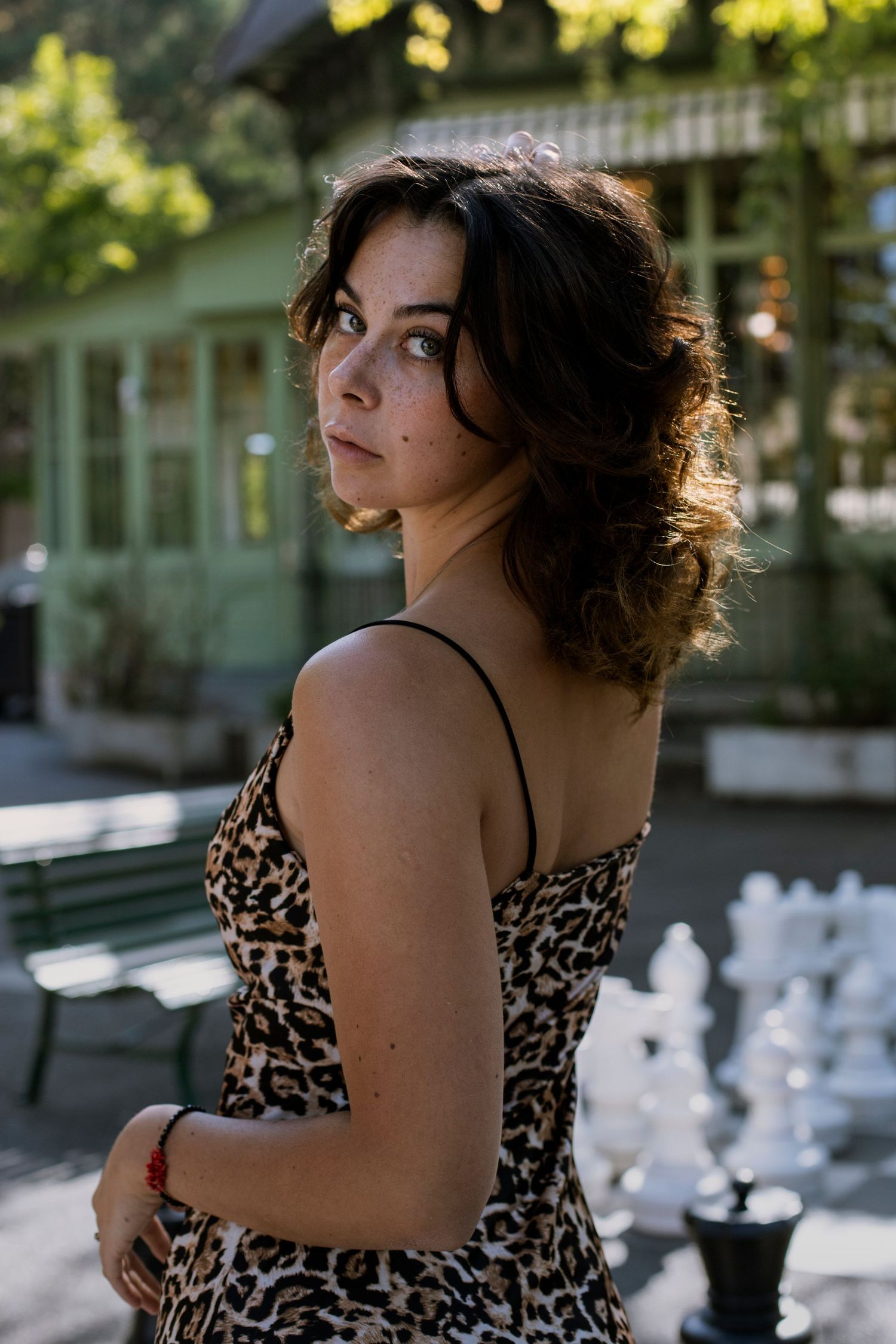 Young woman in leopard print dress looking over her shoulder near a chess set in a sunny park.