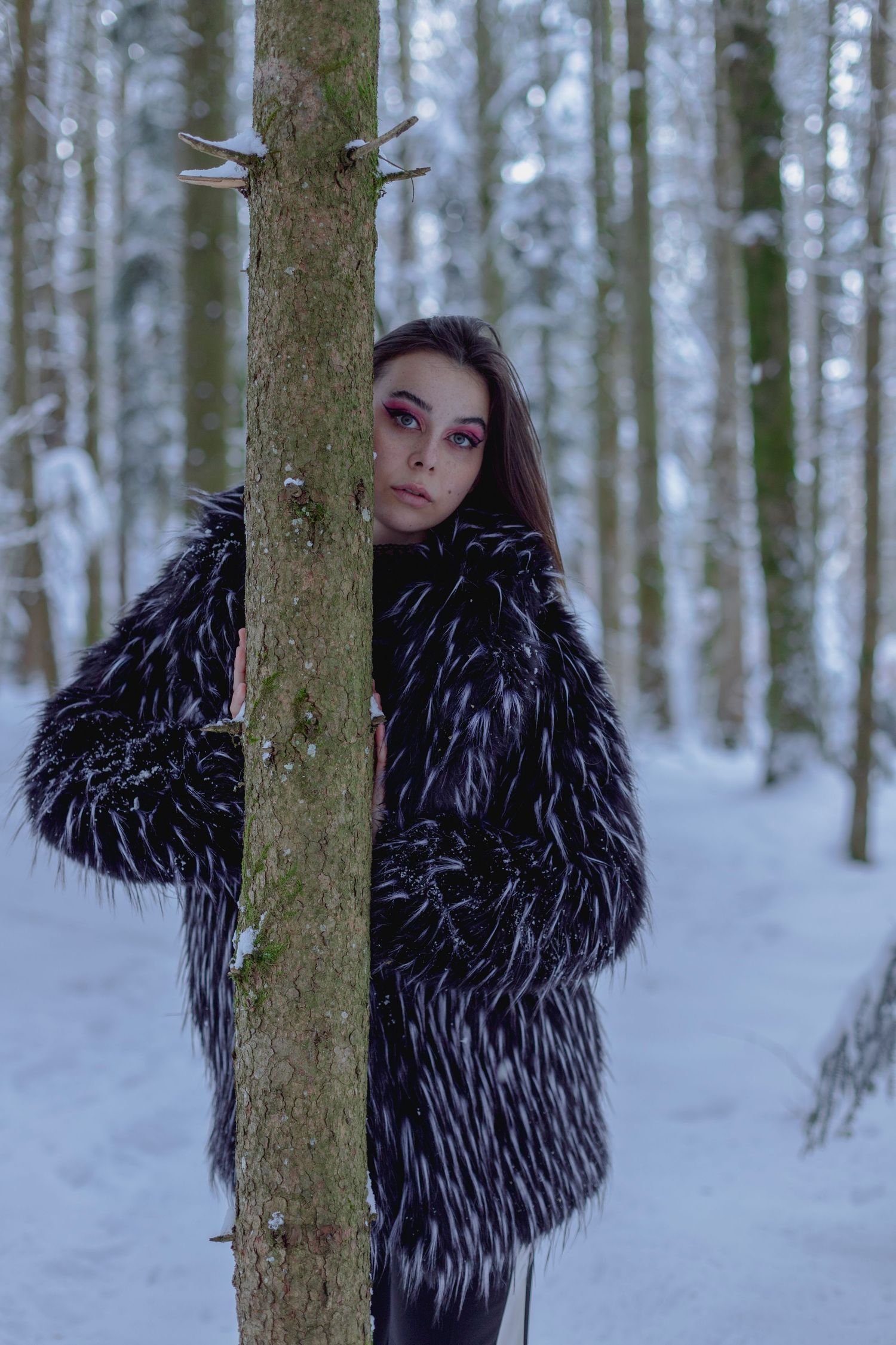 Young woman in black and white faux fur coat posing behind a tree trunk in a snowy forest.