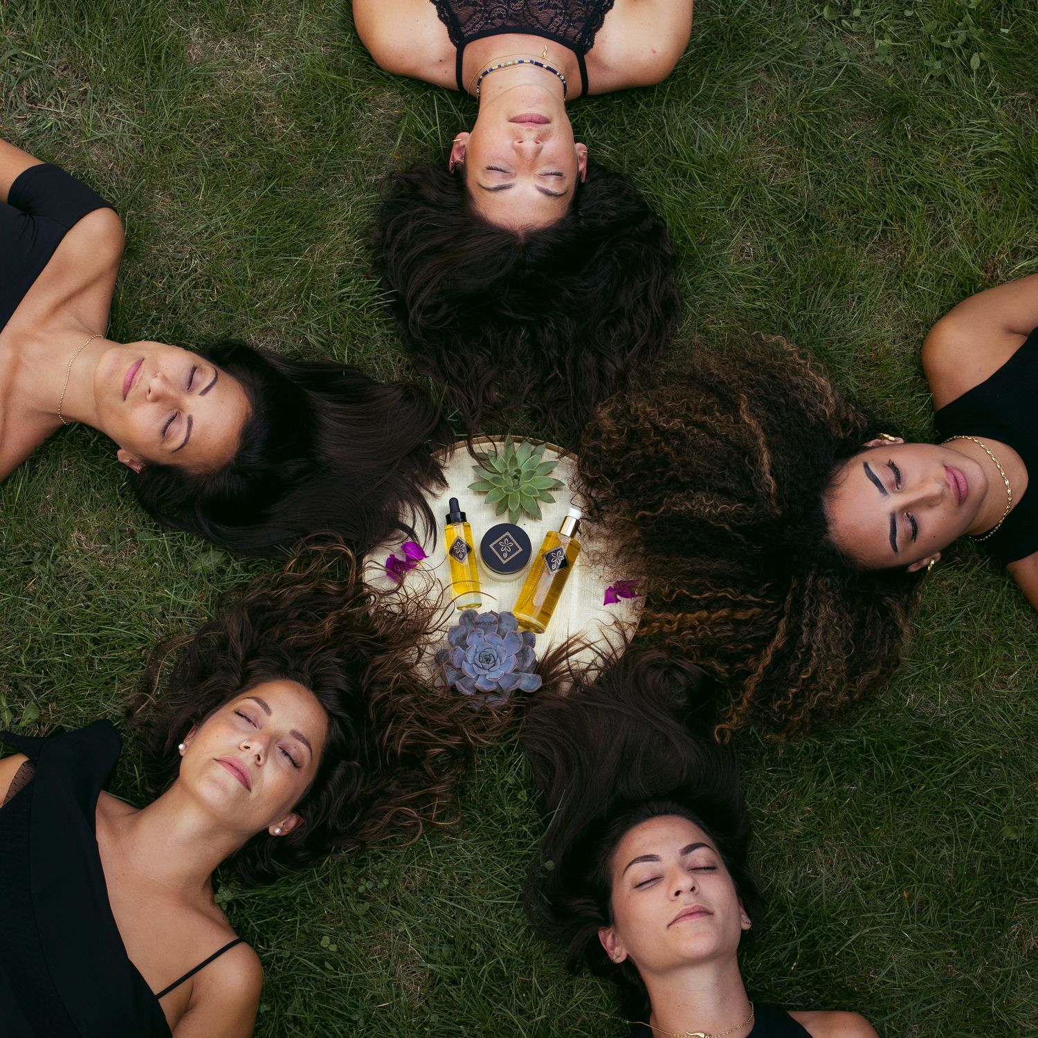 Five women lying on grass in circle with beauty oil products in center.