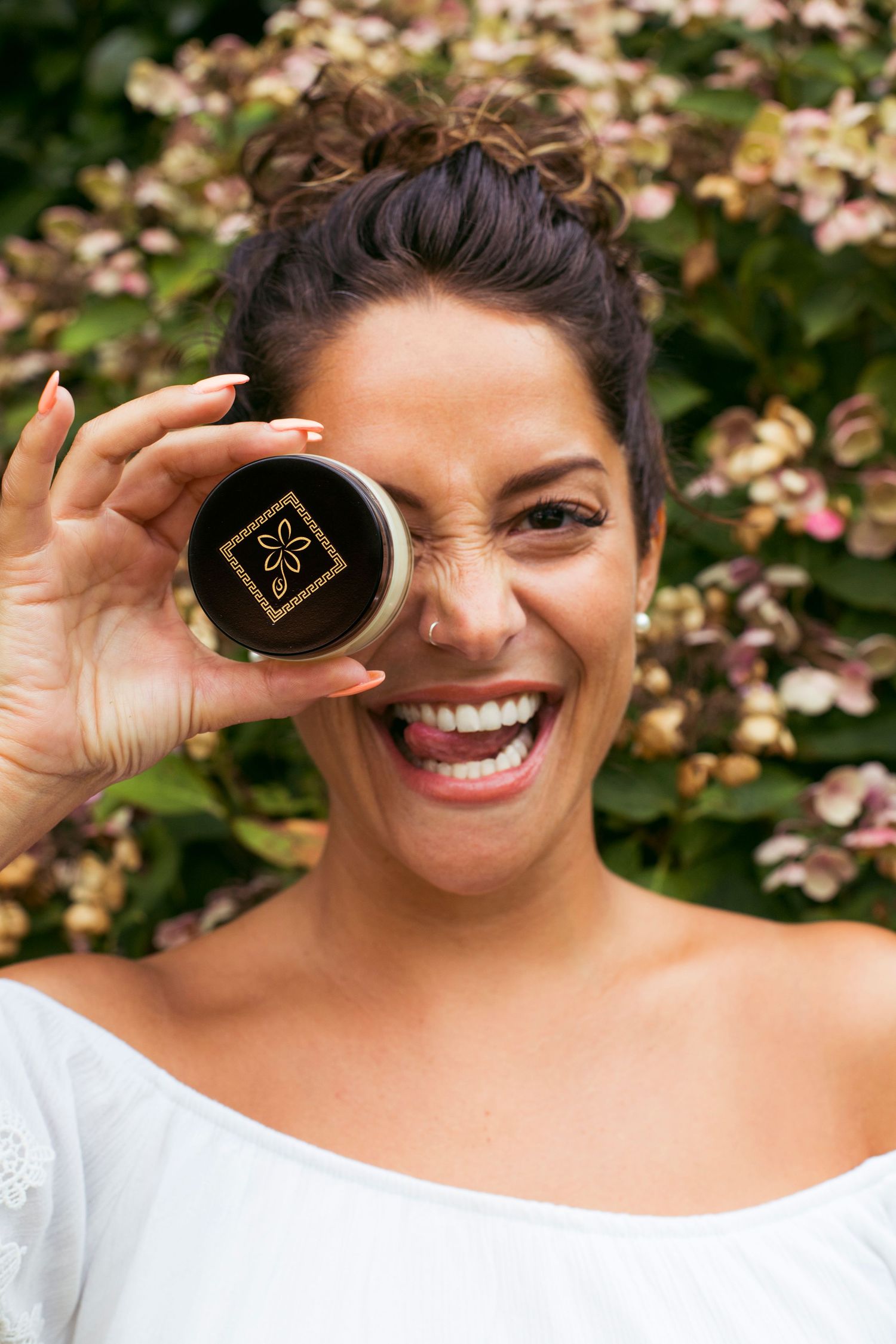 Smiling woman holds black and gold cosmetic jar over her eye outdoors near blooming flowers.