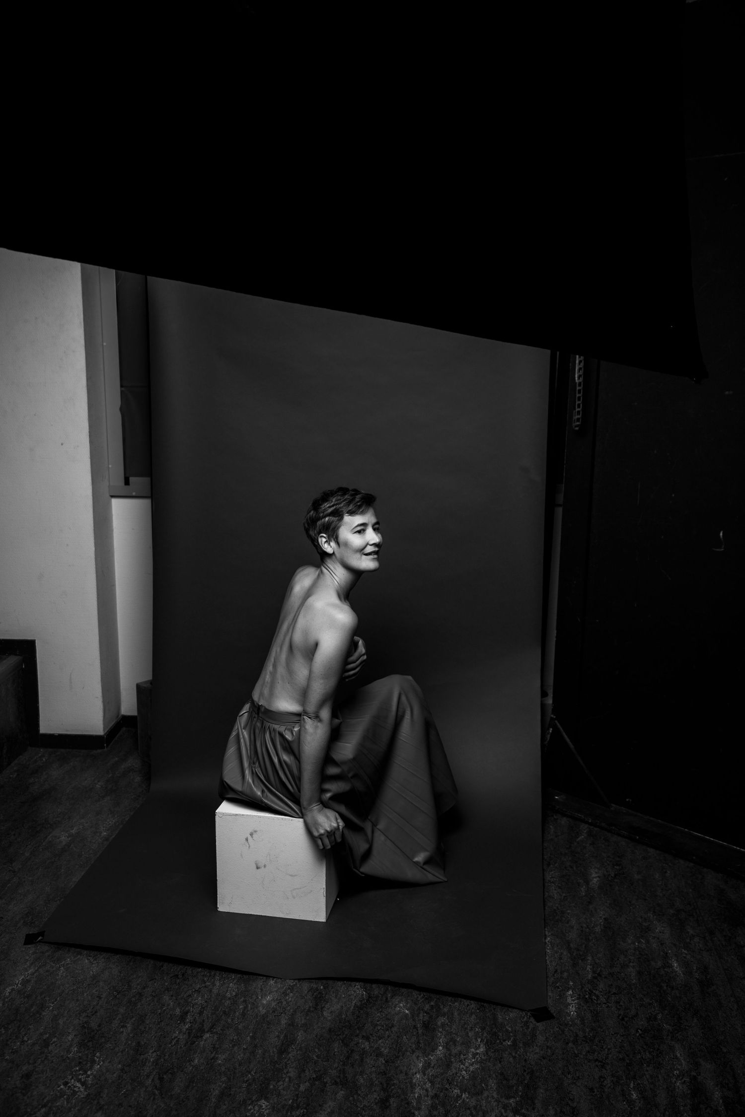 Black and white behind-the-scenes studio shot of topless model laughing on dark backdrop.