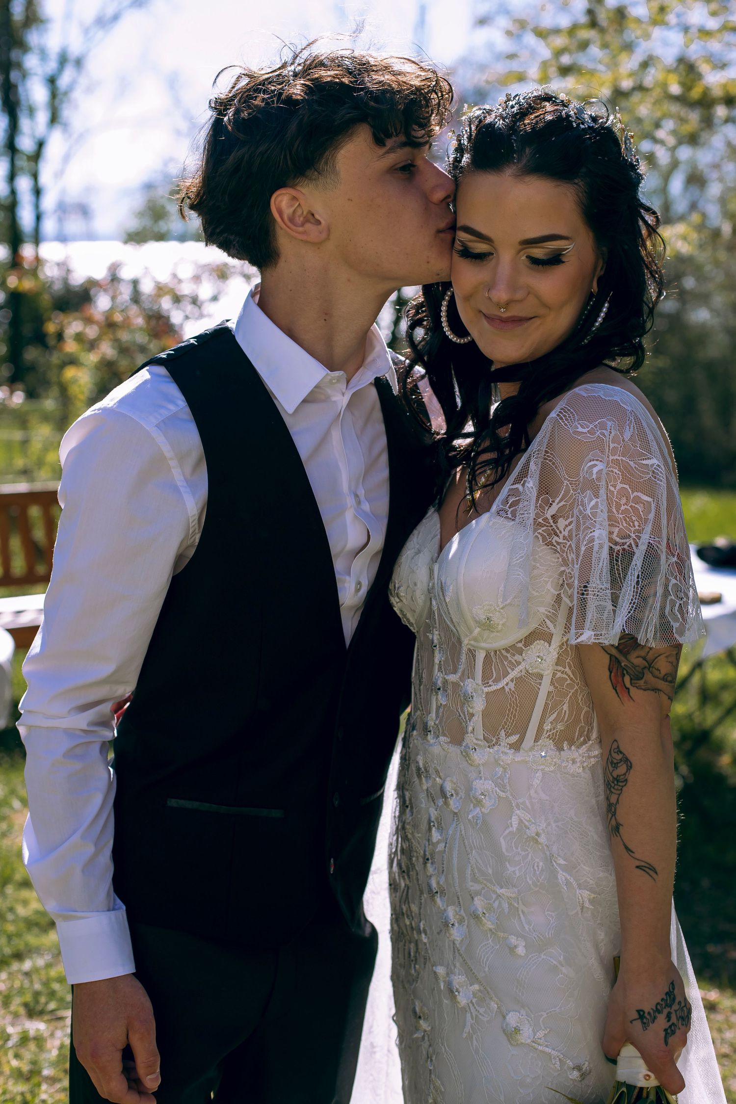 Groom in black vest kissing bride on forehead while she smiles in white lace wedding dress outdoors.