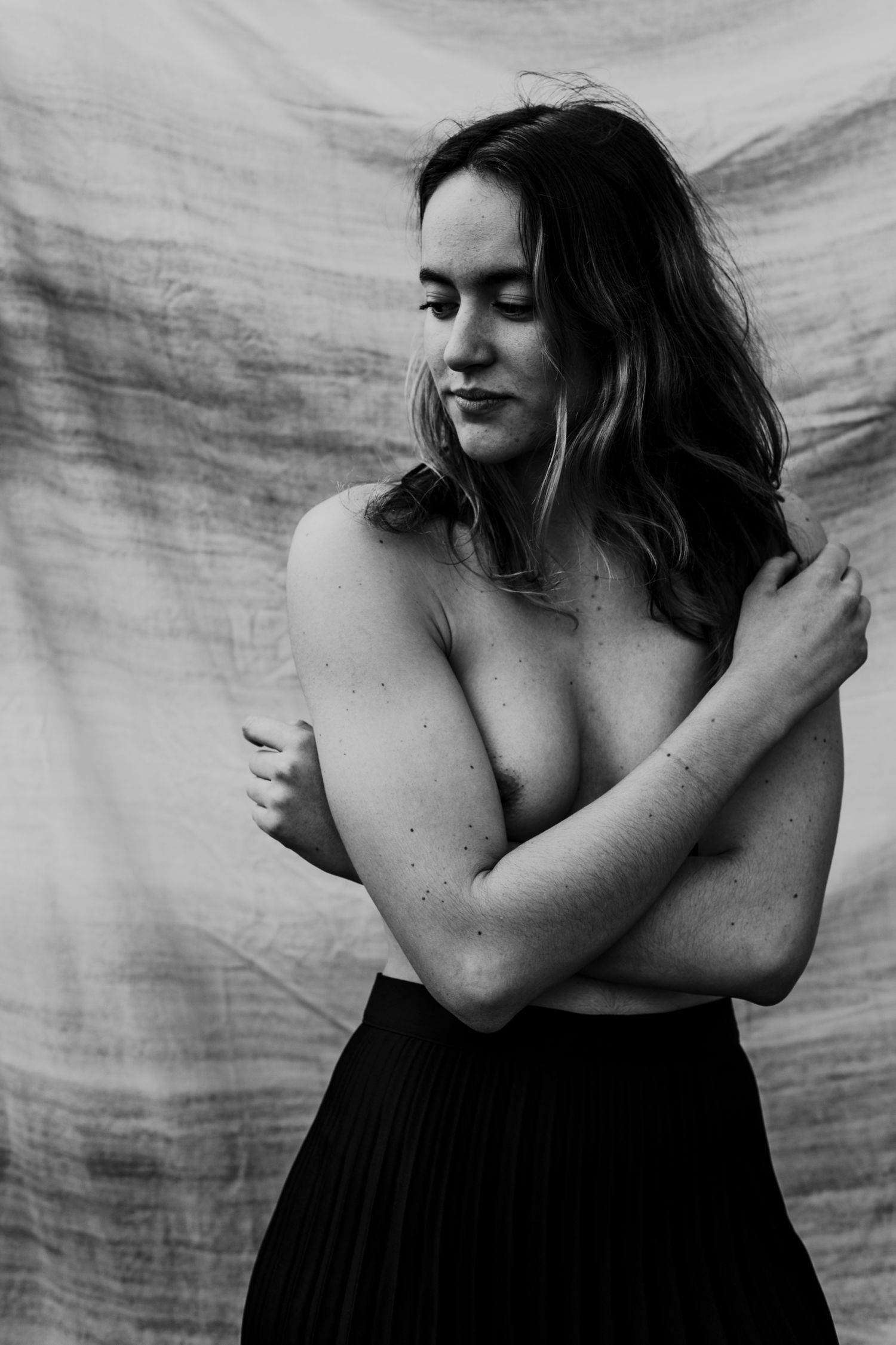Black and white portrait of a smiling topless woman hugging herself against a draped fabric background.