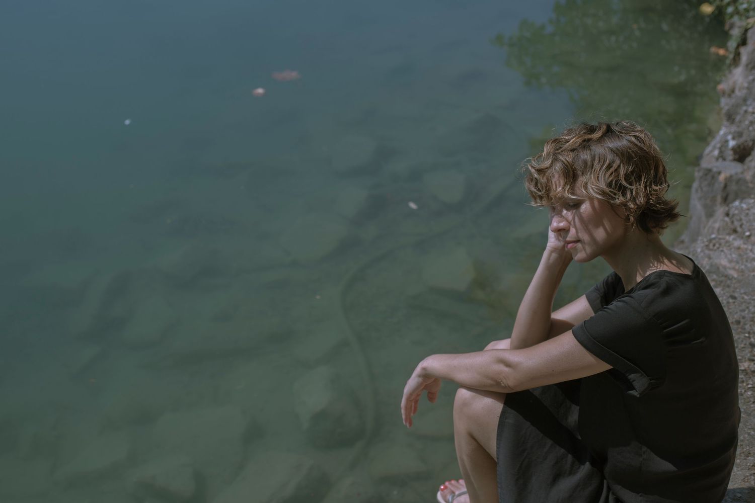 Young person with curly hair sitting pensively on rocks beside a calm green lake in a black t-shirt.