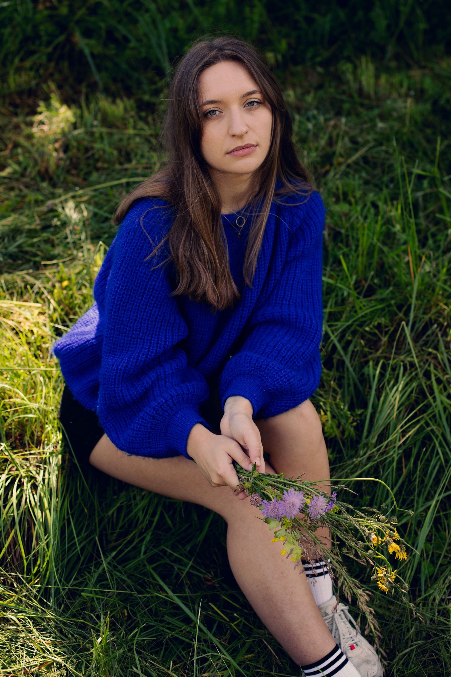 Young woman in blue oversized knit sweater sits on grass holding wildflowers in a field.