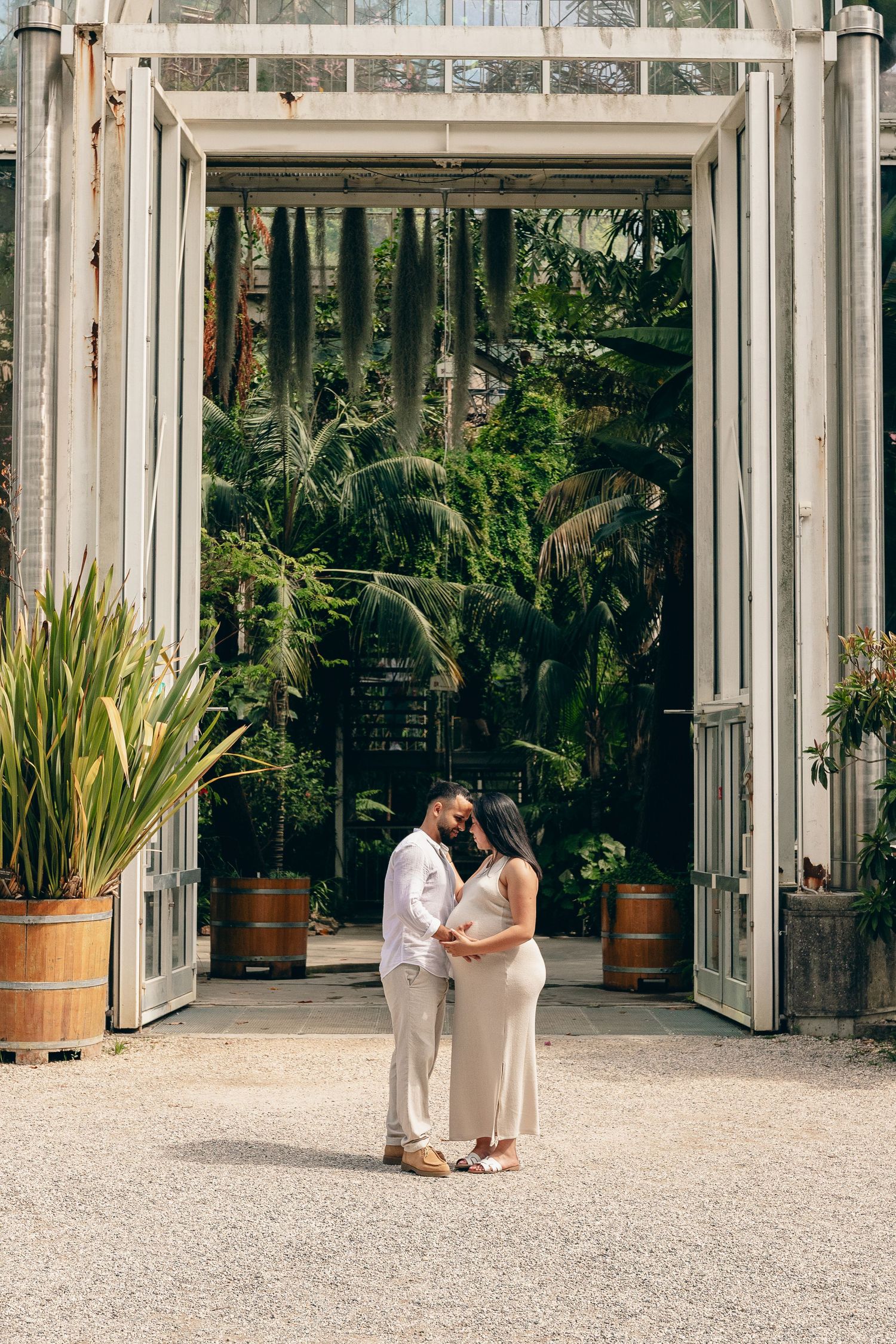 Pregnant couple standing close together in front of a large tropical greenhouse entrance.