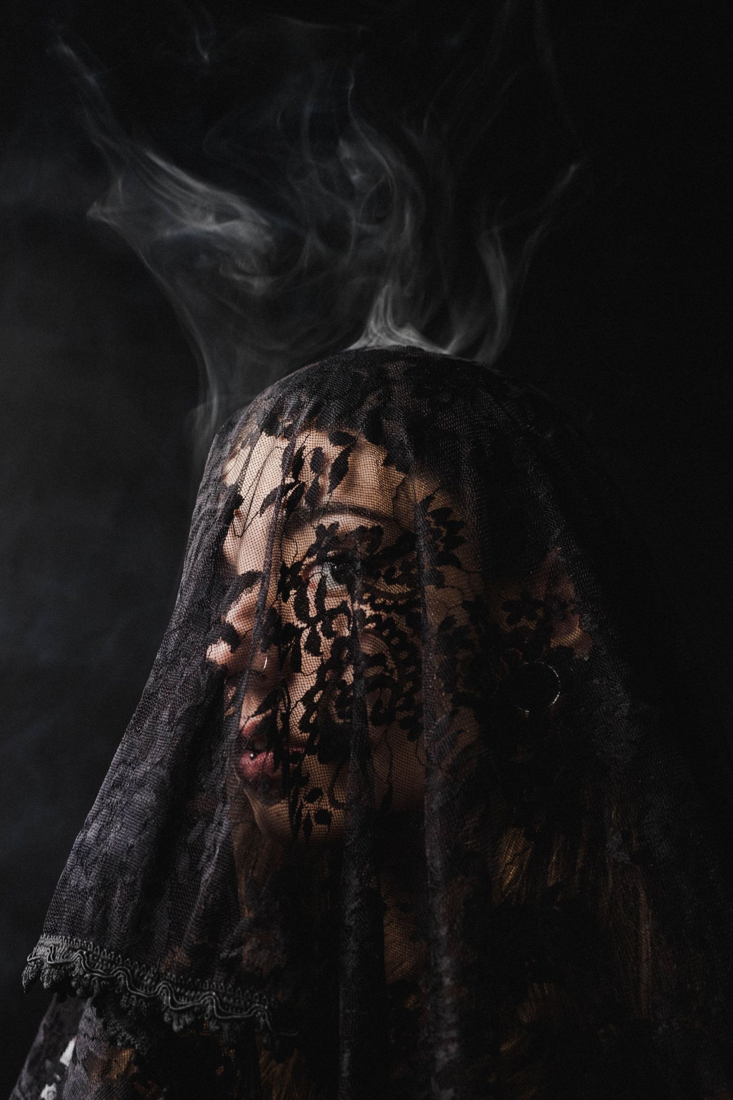Person draped in black floral lace veil with dramatic smoke rising against dark background.