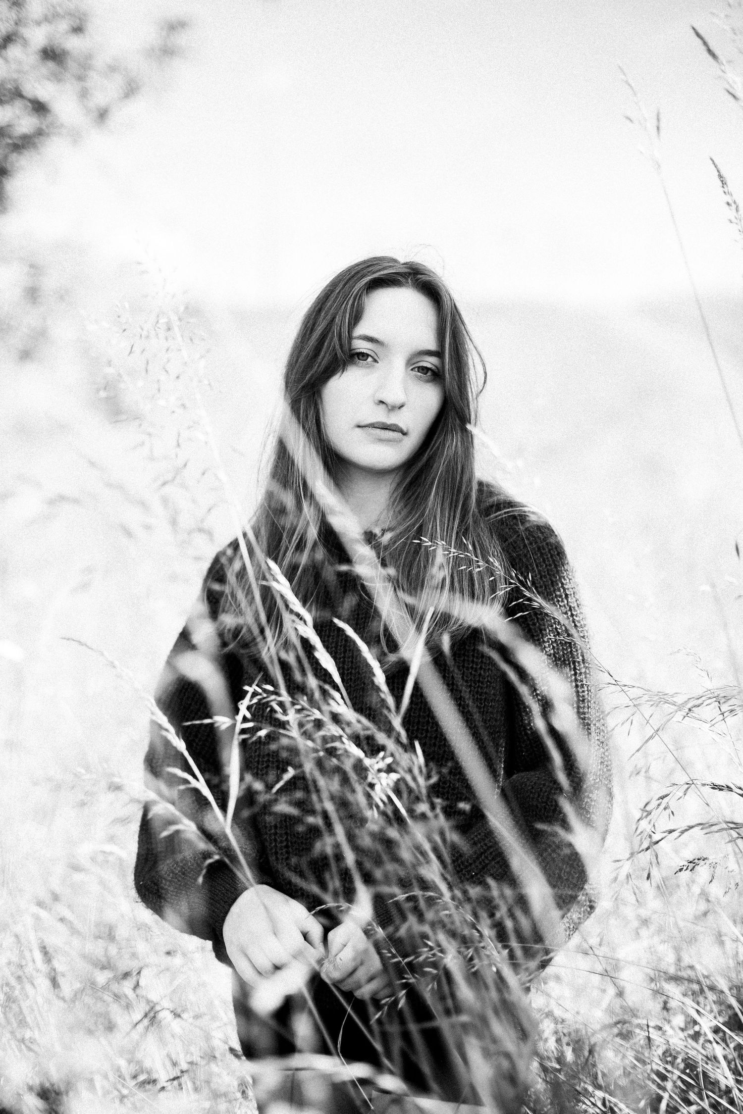 Black and white portrait of young woman with long hair sitting among tall wild grass outdoors.