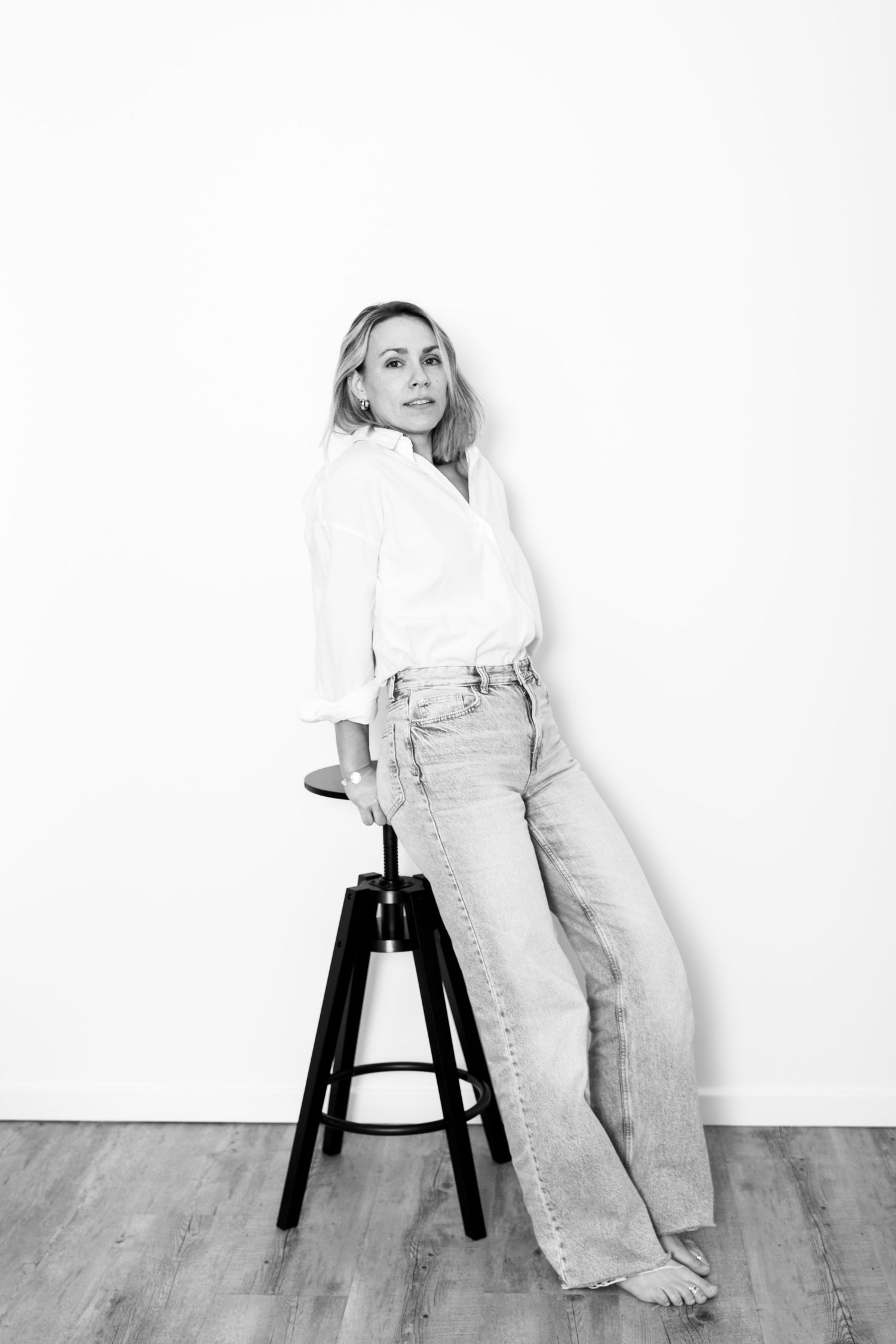Blonde woman in white shirt and jeans leaning on black bar stool, monochrome portrait.