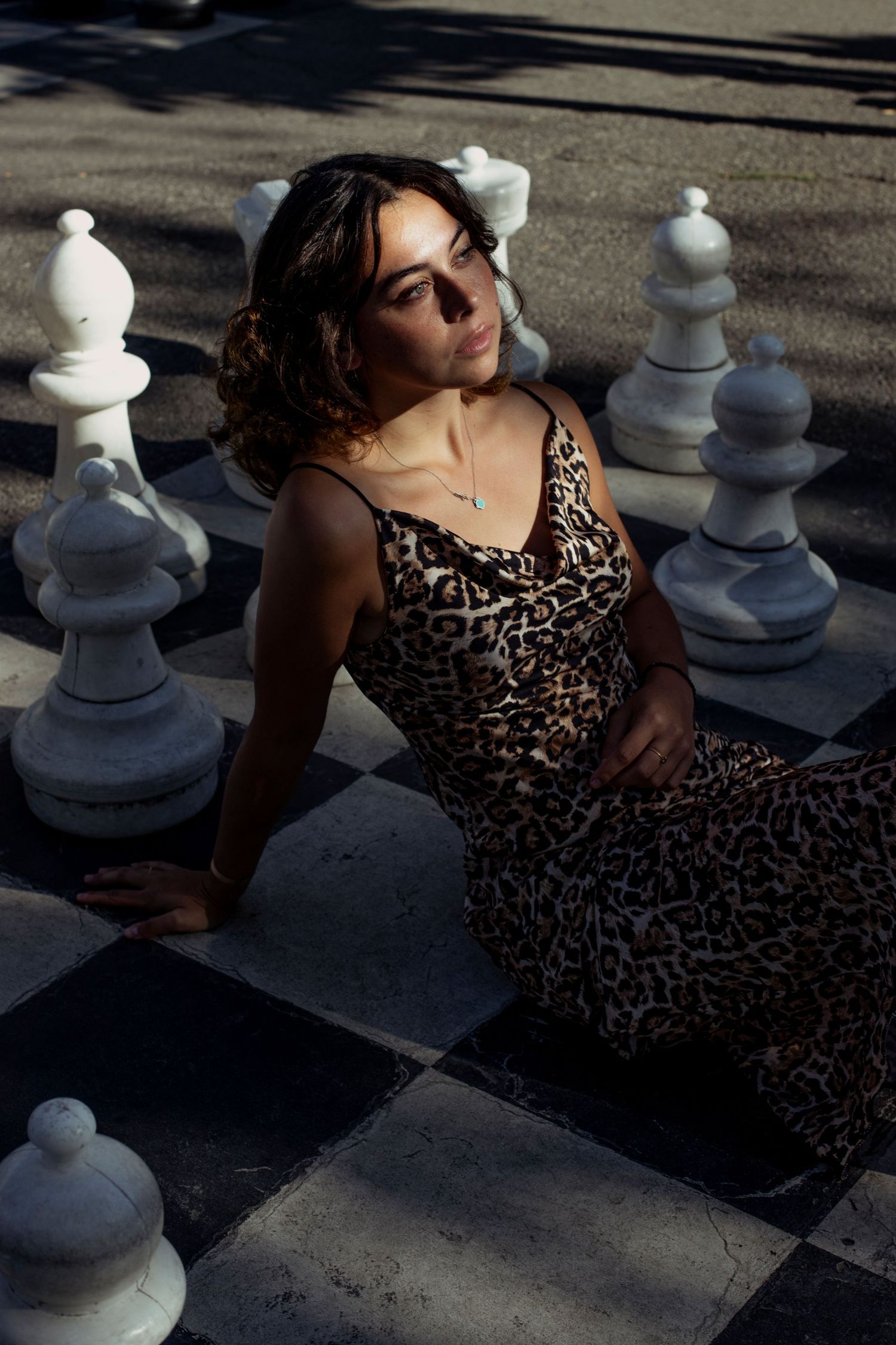 Woman in leopard print dress sitting on giant outdoor chess board surrounded by large pieces.
