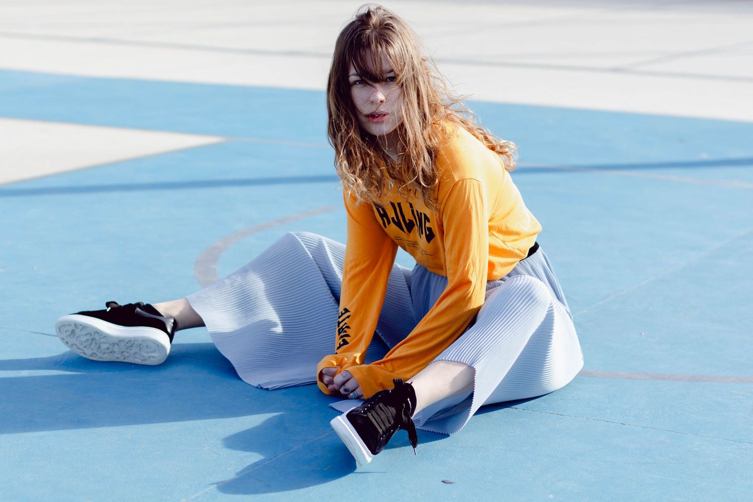Woman in yellow hoodie and white skirt sitting on a blue surface outdoors.