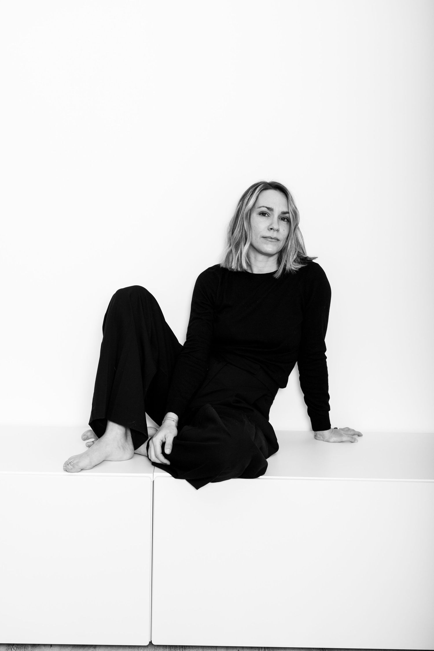 Black and white portrait of blonde woman in black outfit sitting on white surface.