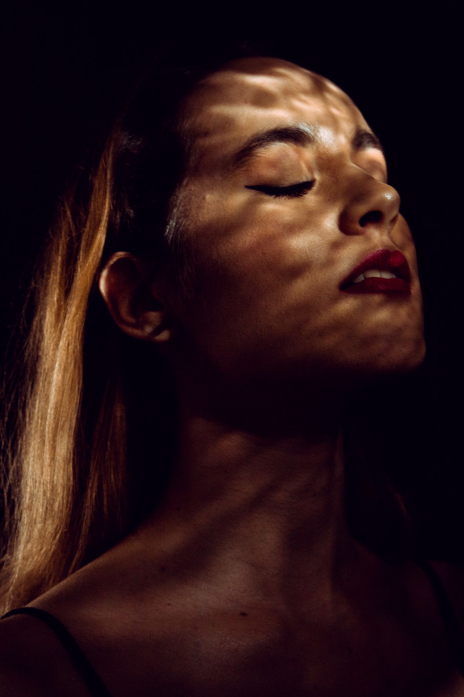 Close-up portrait of woman with red lips and closed eyes with dramatic light pattern on dark background.