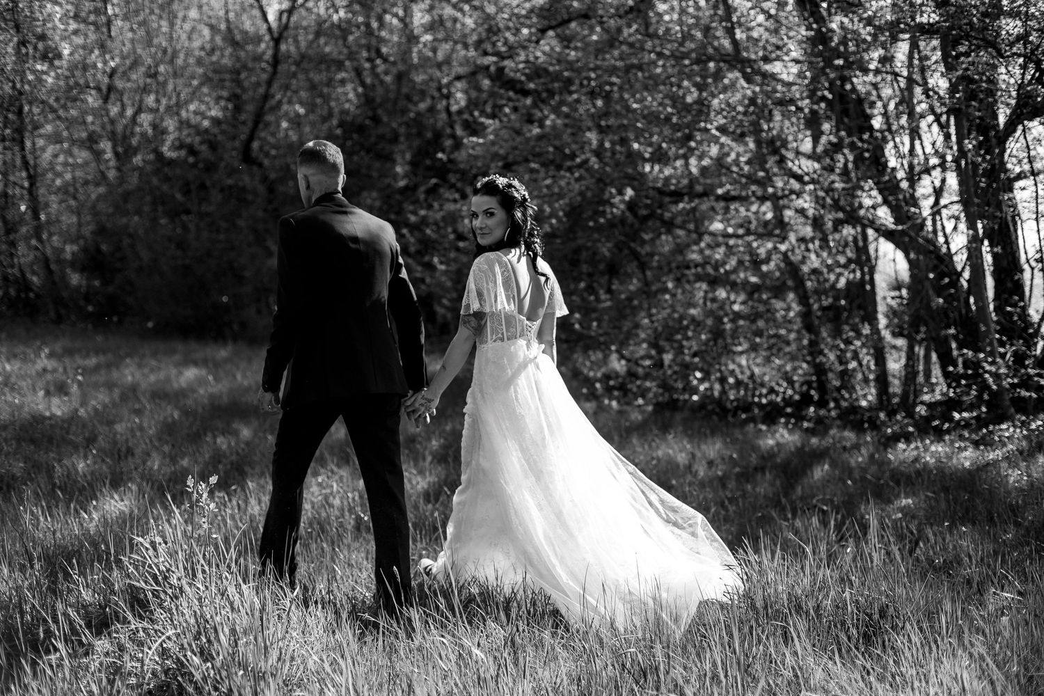 Black and white photo of bride and groom walking hand in hand through a lush green meadow near trees.