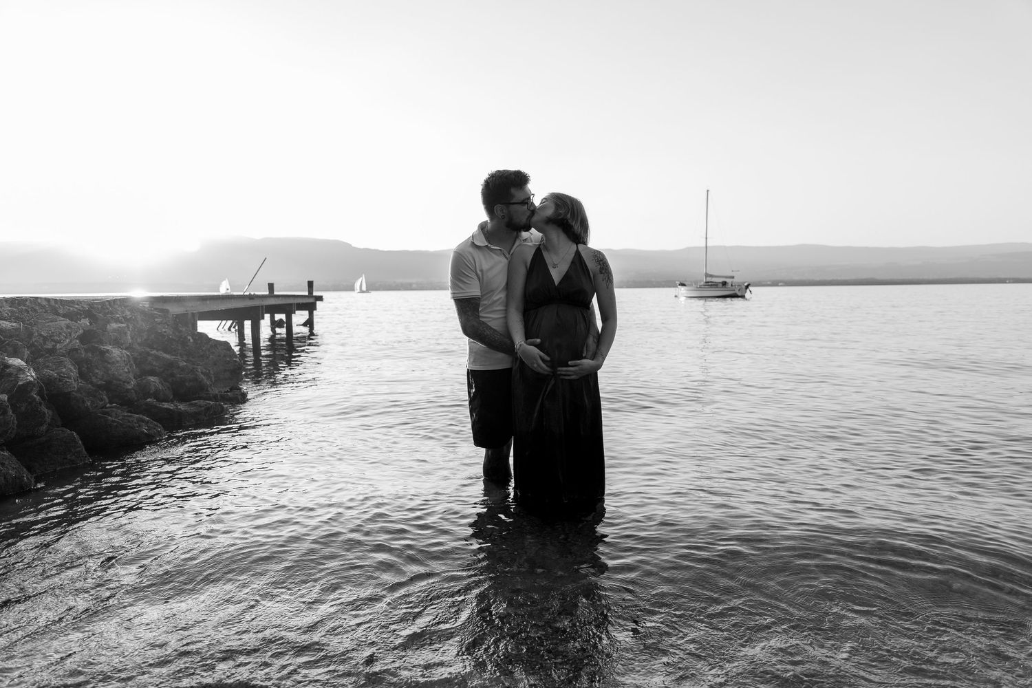 Black and white photo of a couple standing in shallow water at sunset by a pier.