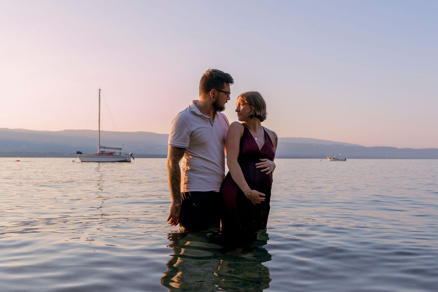 Pregnant couple standing in a calm lake at sunset with a sailboat in the background.