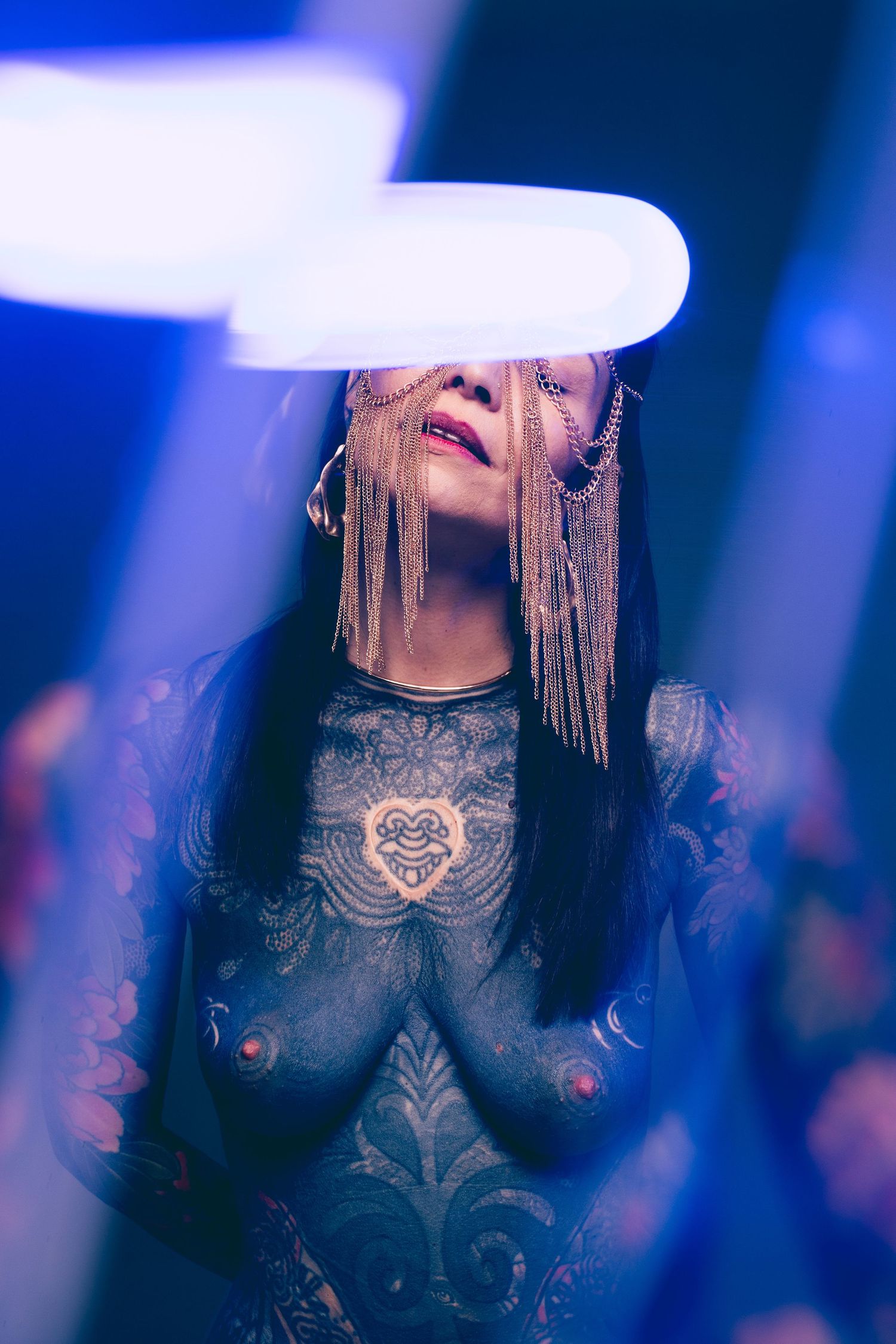 Heavily tattooed woman in sheer bodysuit looks upward under blue neon lighting with gold chain headpiece.
