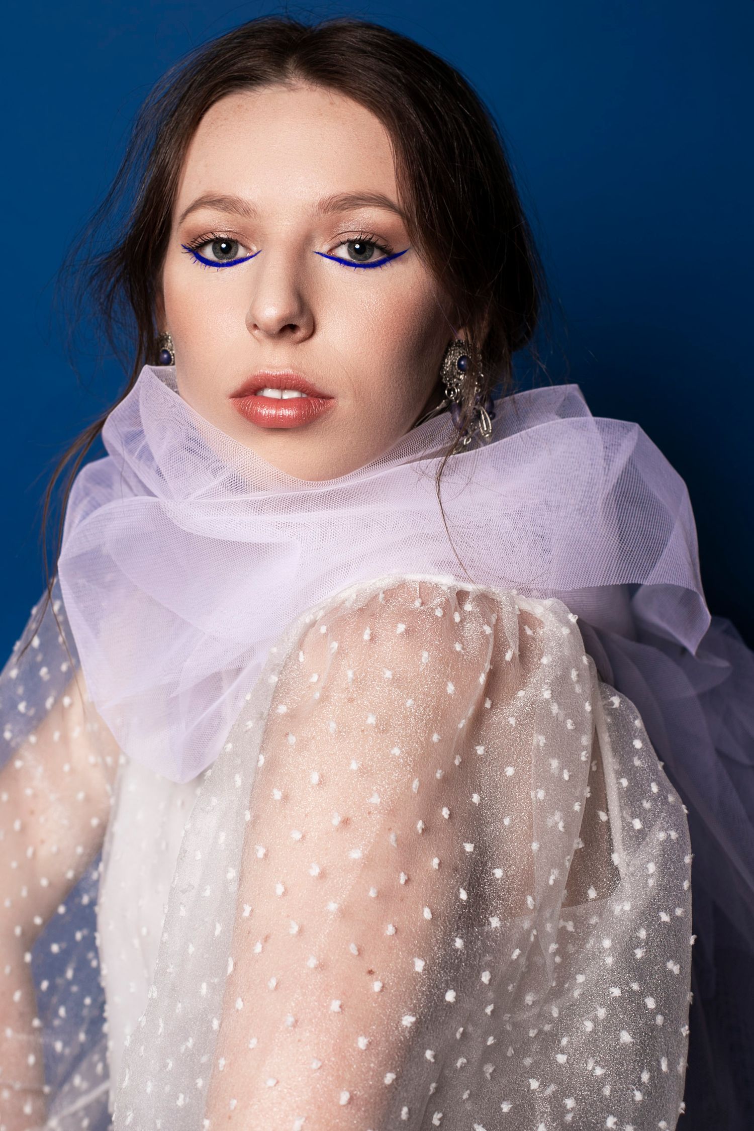 Woman with bold blue eyeliner wearing a sheer white polka dot veil against a blue background.