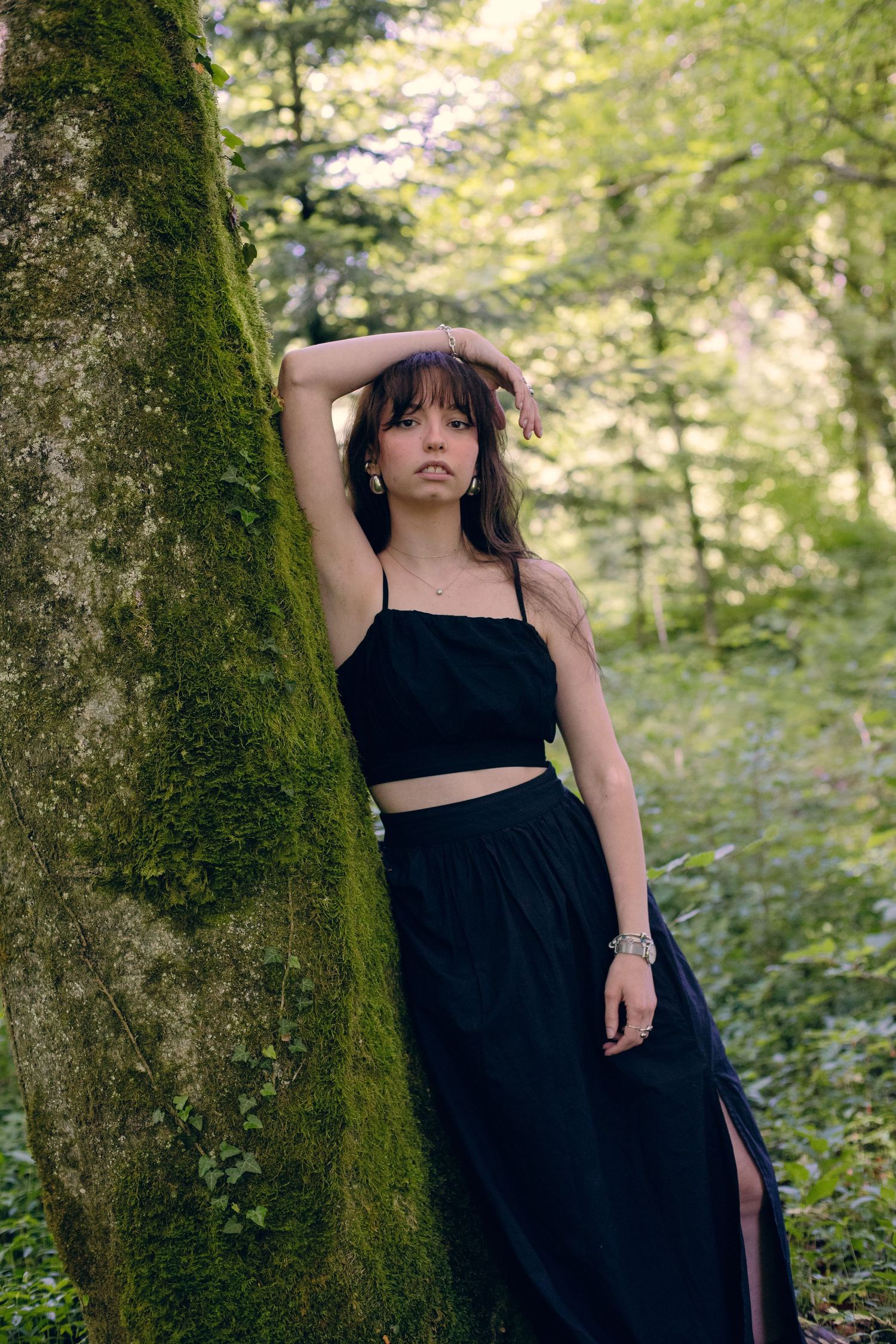 Young woman in black crop top and maxi skirt leaning against moss-covered tree in lush green forest.