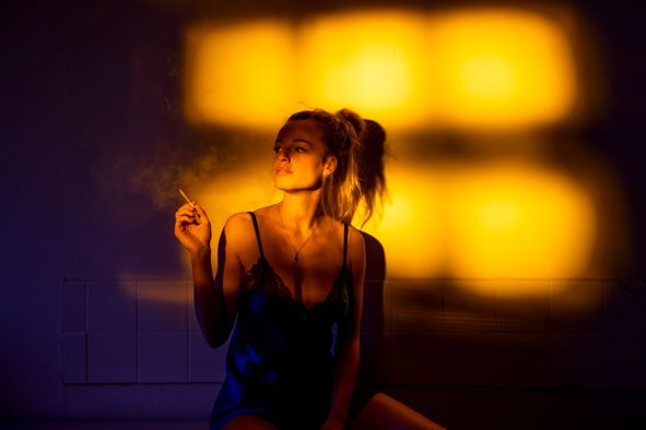 Woman in black bodysuit smoking a cigarette under warm orange ambient lighting.