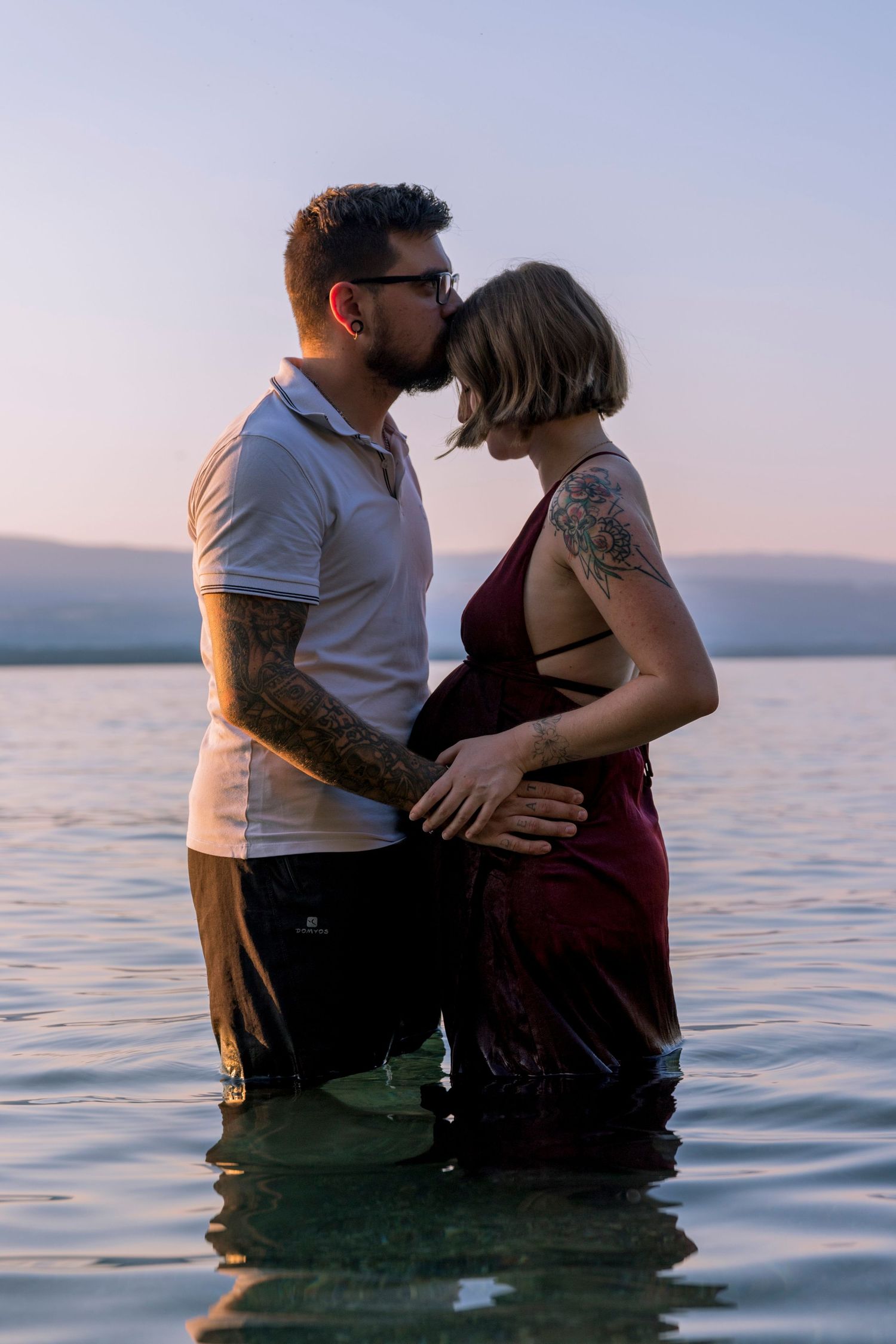 Tattooed couple sharing a kiss while standing in lake water at dusk, woman visibly pregnant.