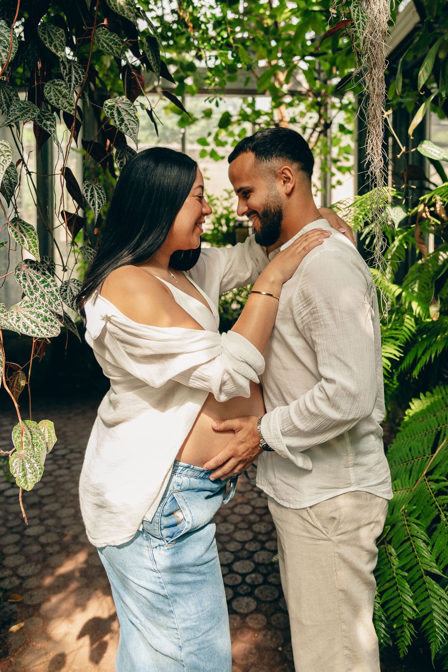 Smiling pregnant couple in white outfits embracing in sunlit tropical greenhouse maternity portrait.